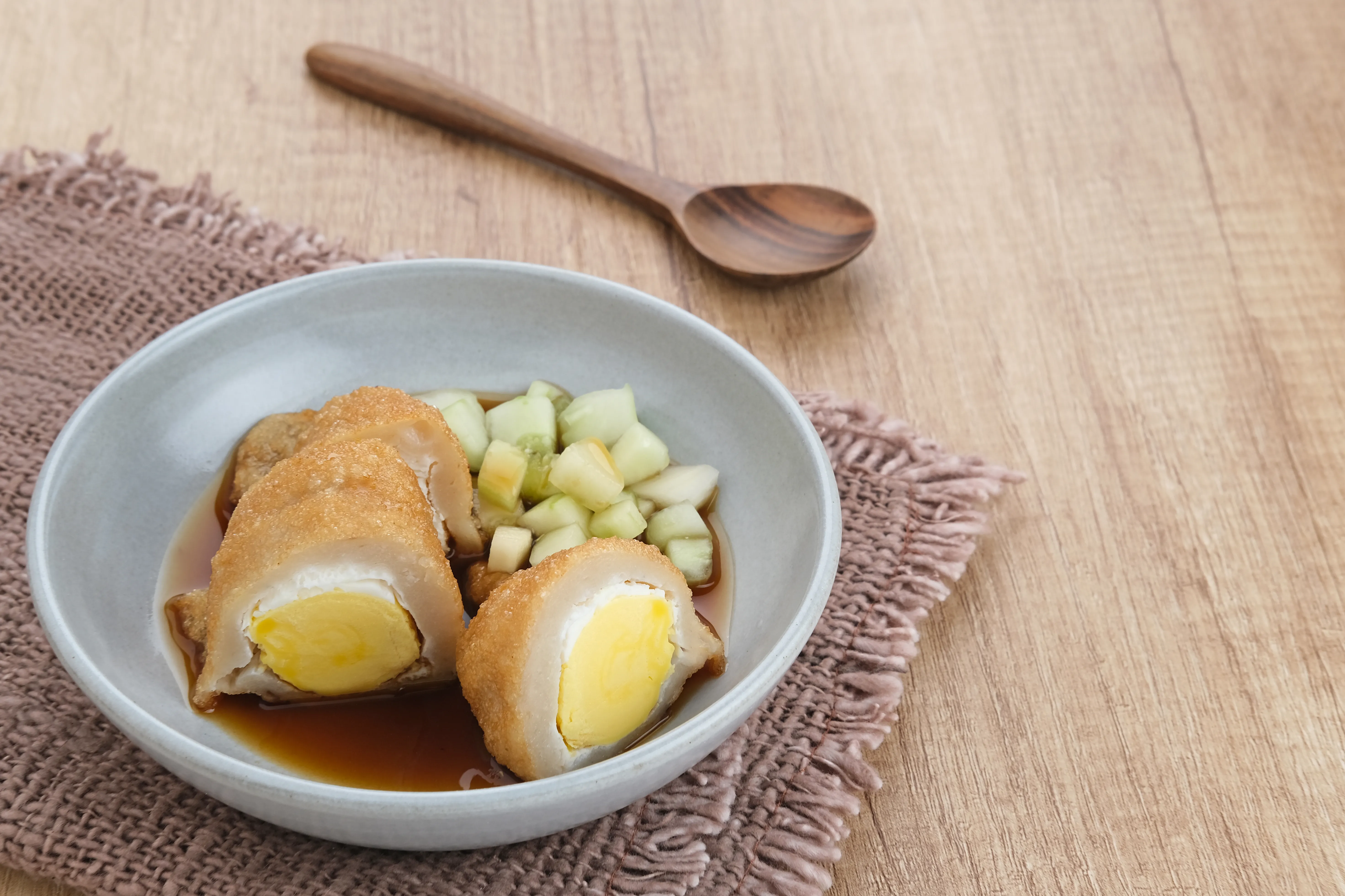 A grey bowl of Pempek Kapal Selam, featuring sliced egg-filled fishcakes and diced cucumber in a dark cuko sauce, set on a wooden table.
