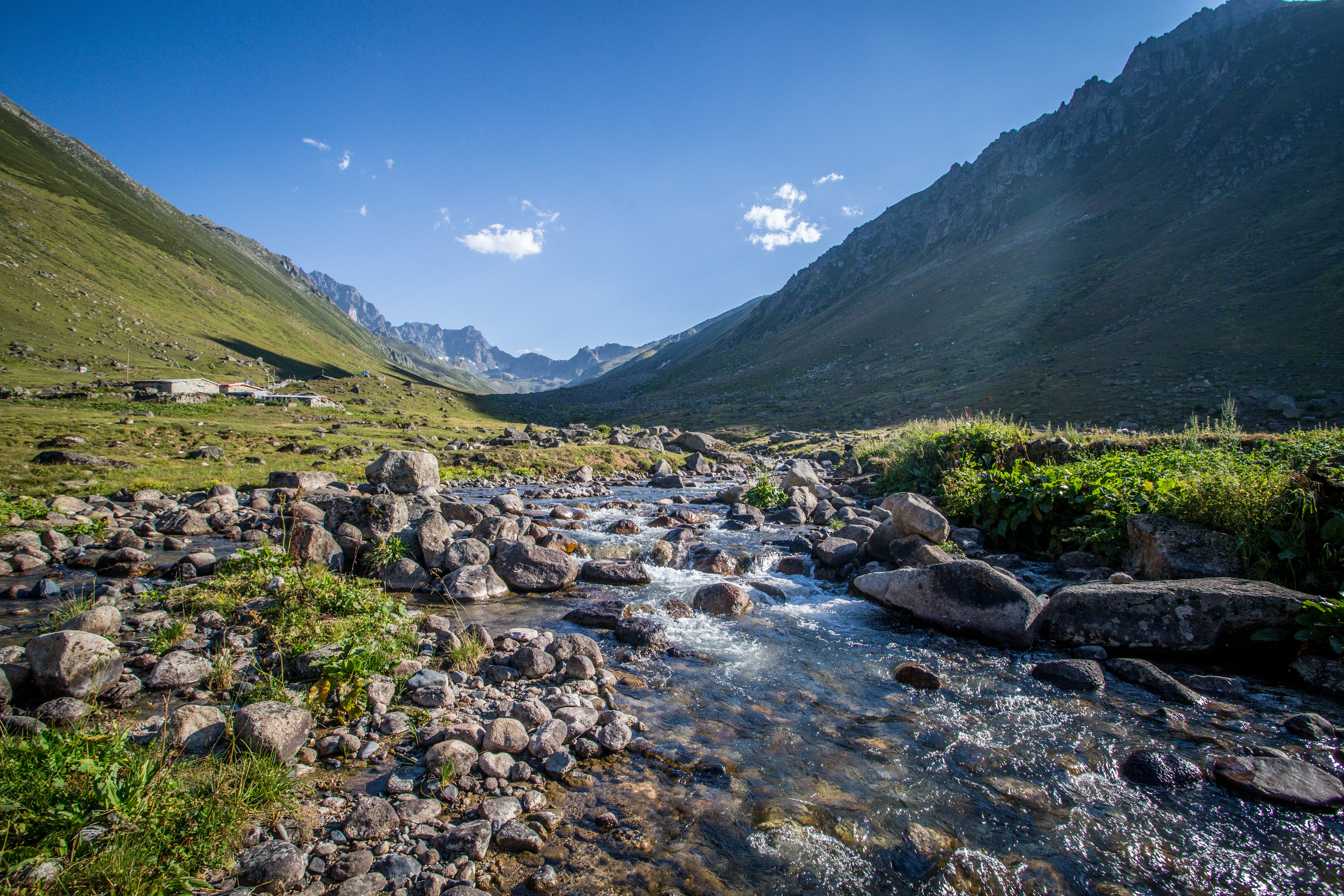 A rocky stream flowing through a lush green mountain valley under a clear blue sky