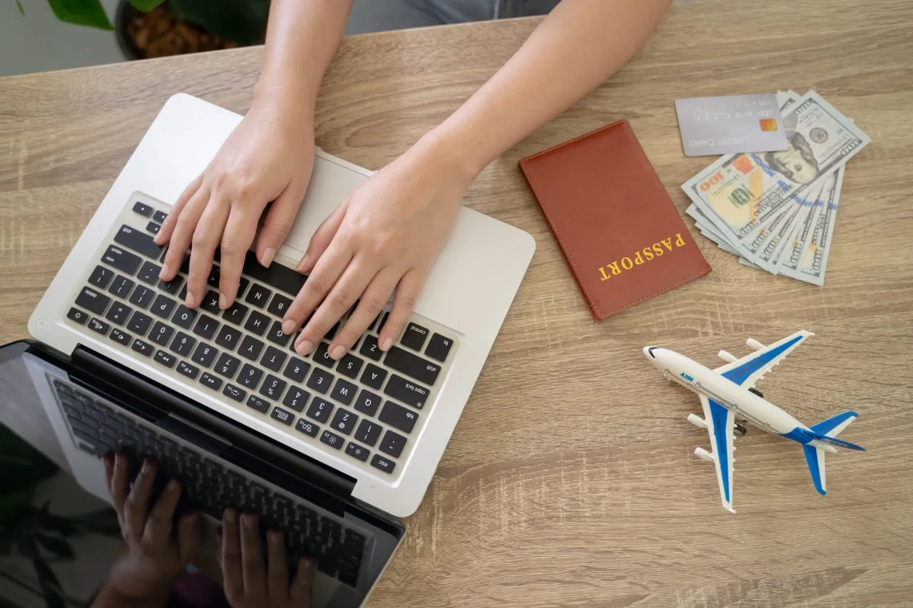 The process of planning a trip, featuring a person using a laptop alongside a passport, credit card, and currency