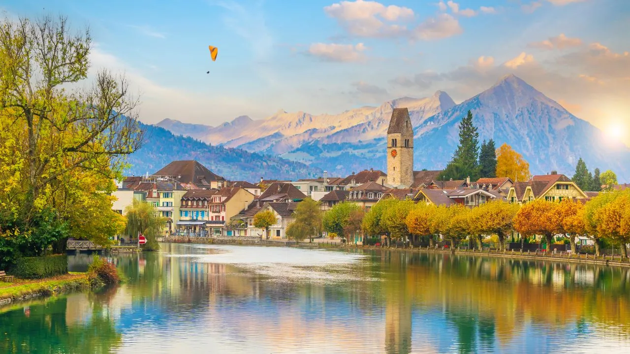 Riverside town with autumn trees, mountains, and a hot air balloon in the sky