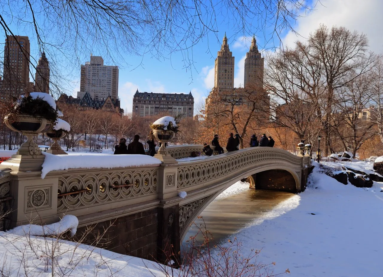 Snow-covered park with a decorative bridge and city skyline in the background