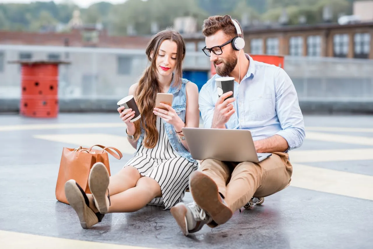 Two young adults sitting outdoors, using smartphones and a laptop while wearing headphones