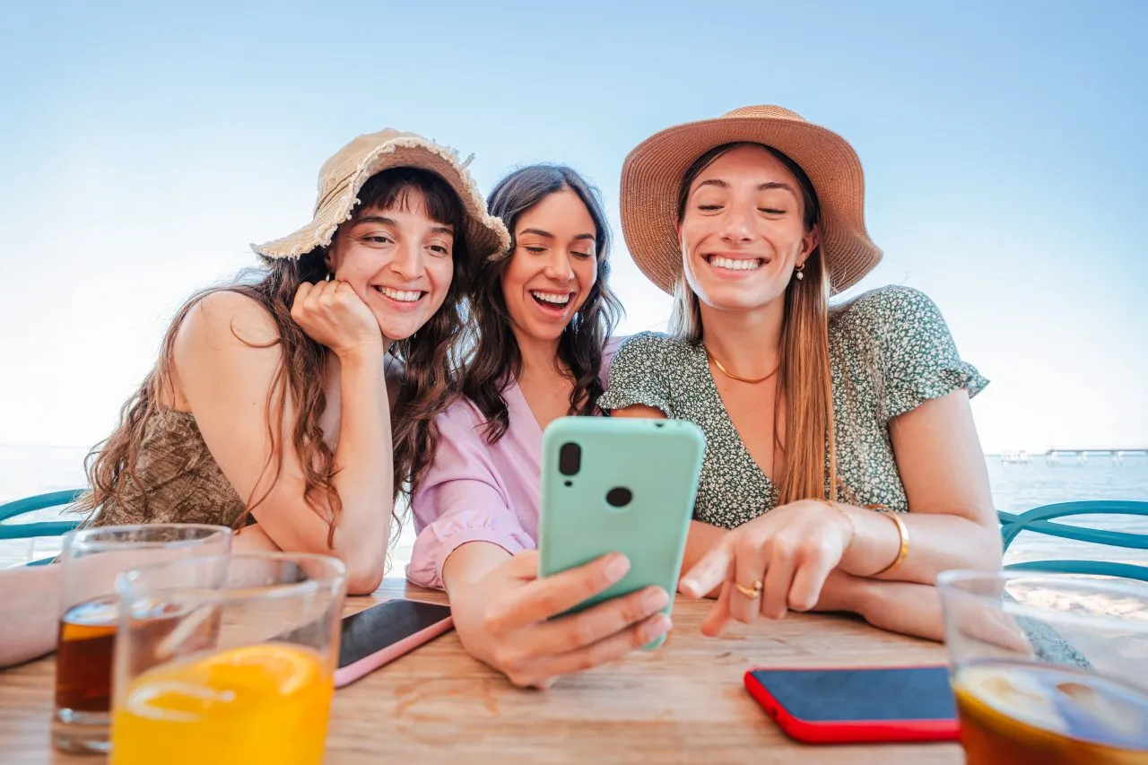 Three friends smiling and taking a selfie at an outdoor table with drinks