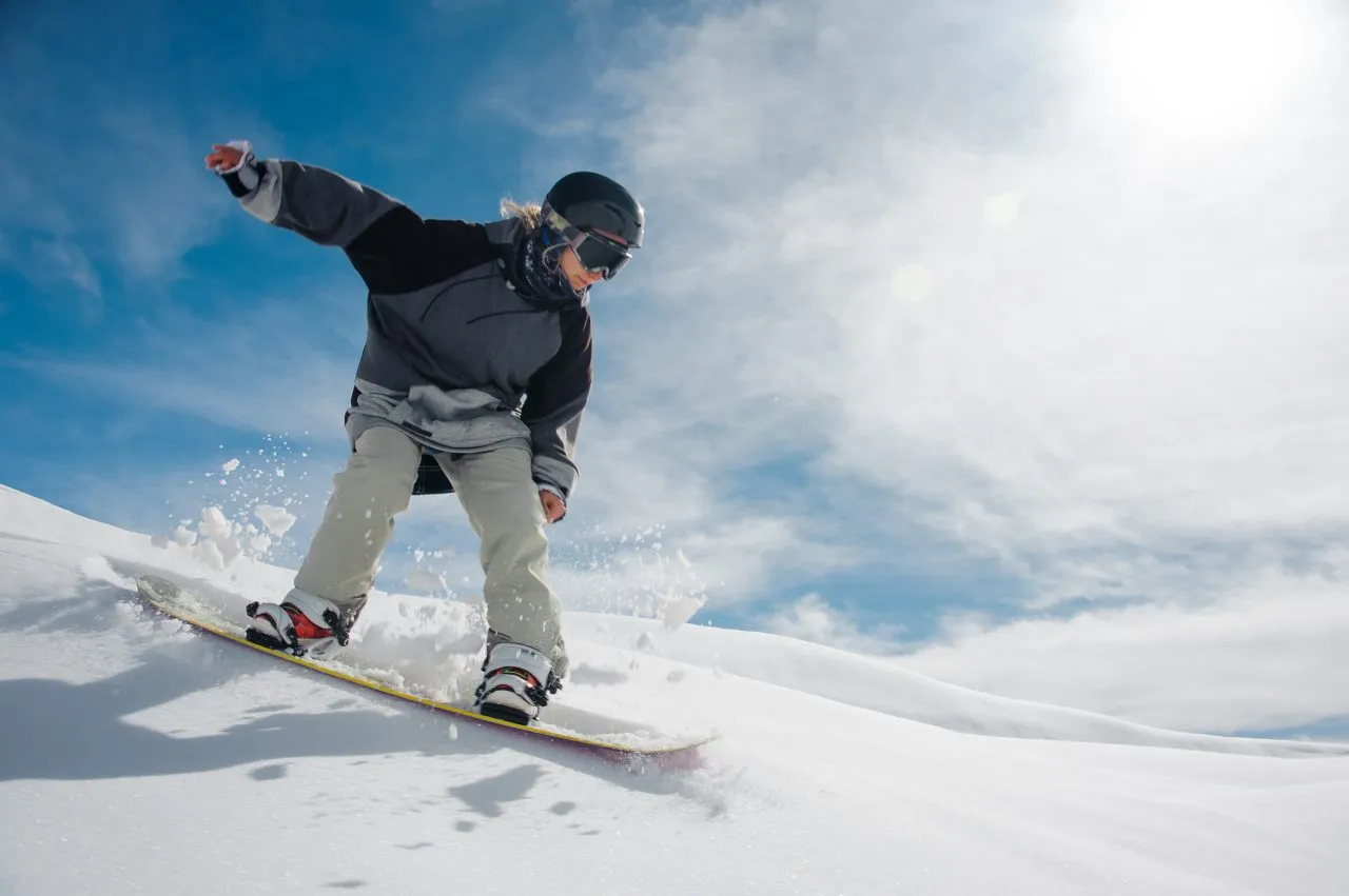Snowboarder riding through fresh powder on a mountain slope under a bright blue winter sky