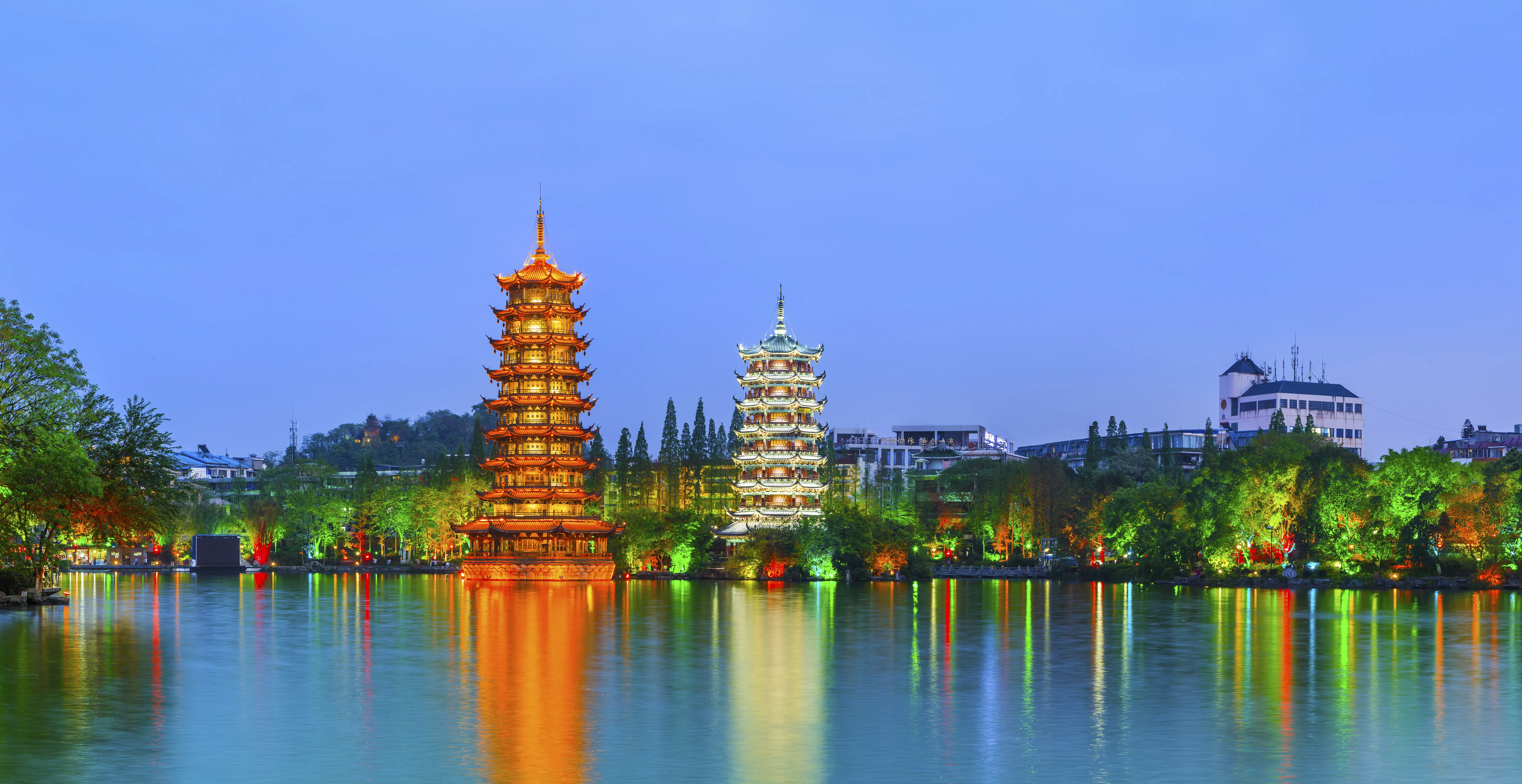 A wide-angle, eye-level shot of the Sun and Moon Twin Pagodas standing illuminated on Shanhu Lake in Guilin, China