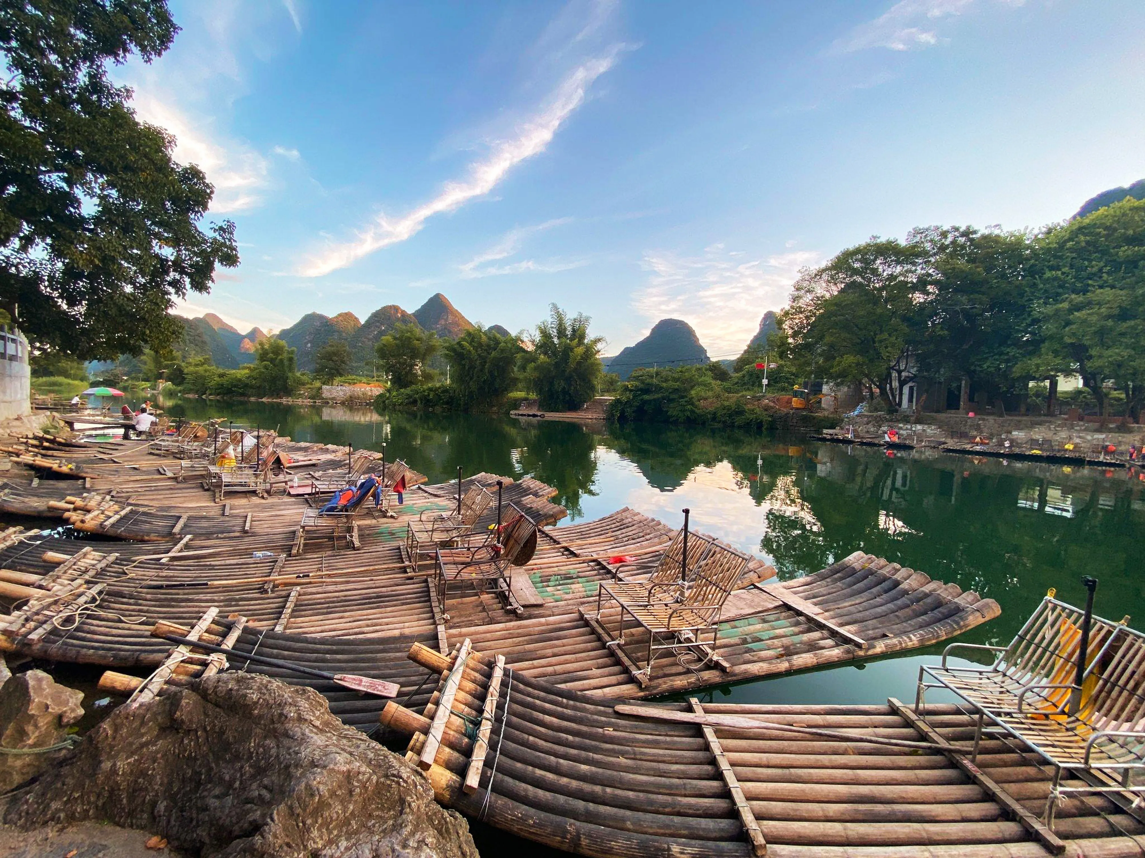 A wide-angle, eye-level shot of numerous traditional bamboo rafts with metal chairs lined up along the calm riverbank of the Yulong River in Yangshuo, China.