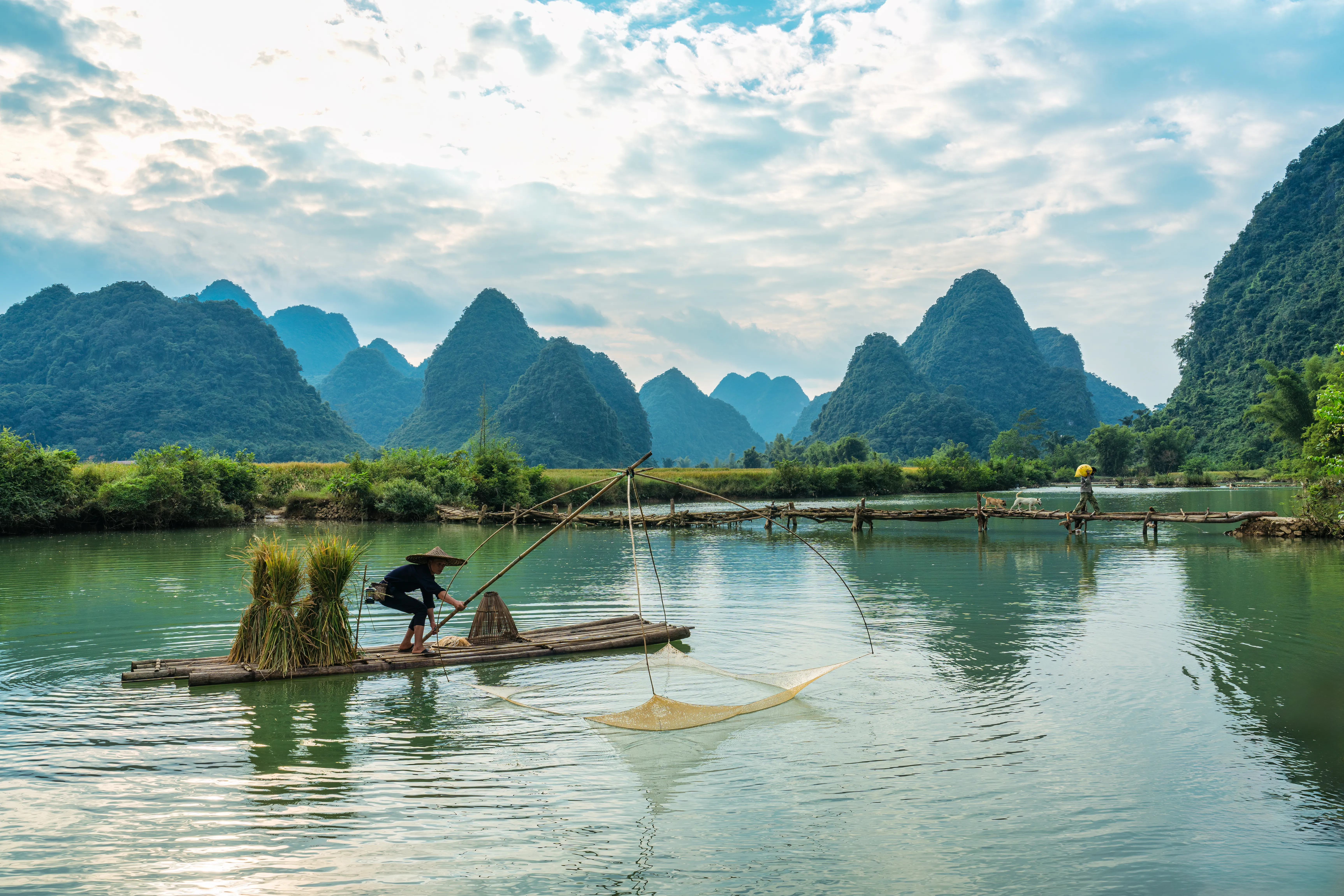 A wide-angle, eye-level shot of a fisherman wearing a traditional conical hat on a bamboo raft, using a large dip net to fish in a calm, green river in Yangshuo, China.