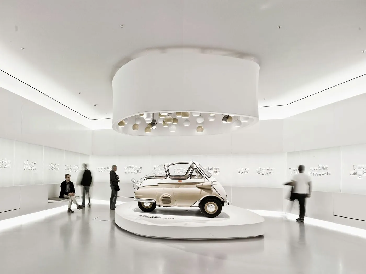 A wide-angle, eye-level shot of a vintage beige BMW Isetta microcar displayed on a raised oval platform inside a sleek, minimalist white museum gallery.