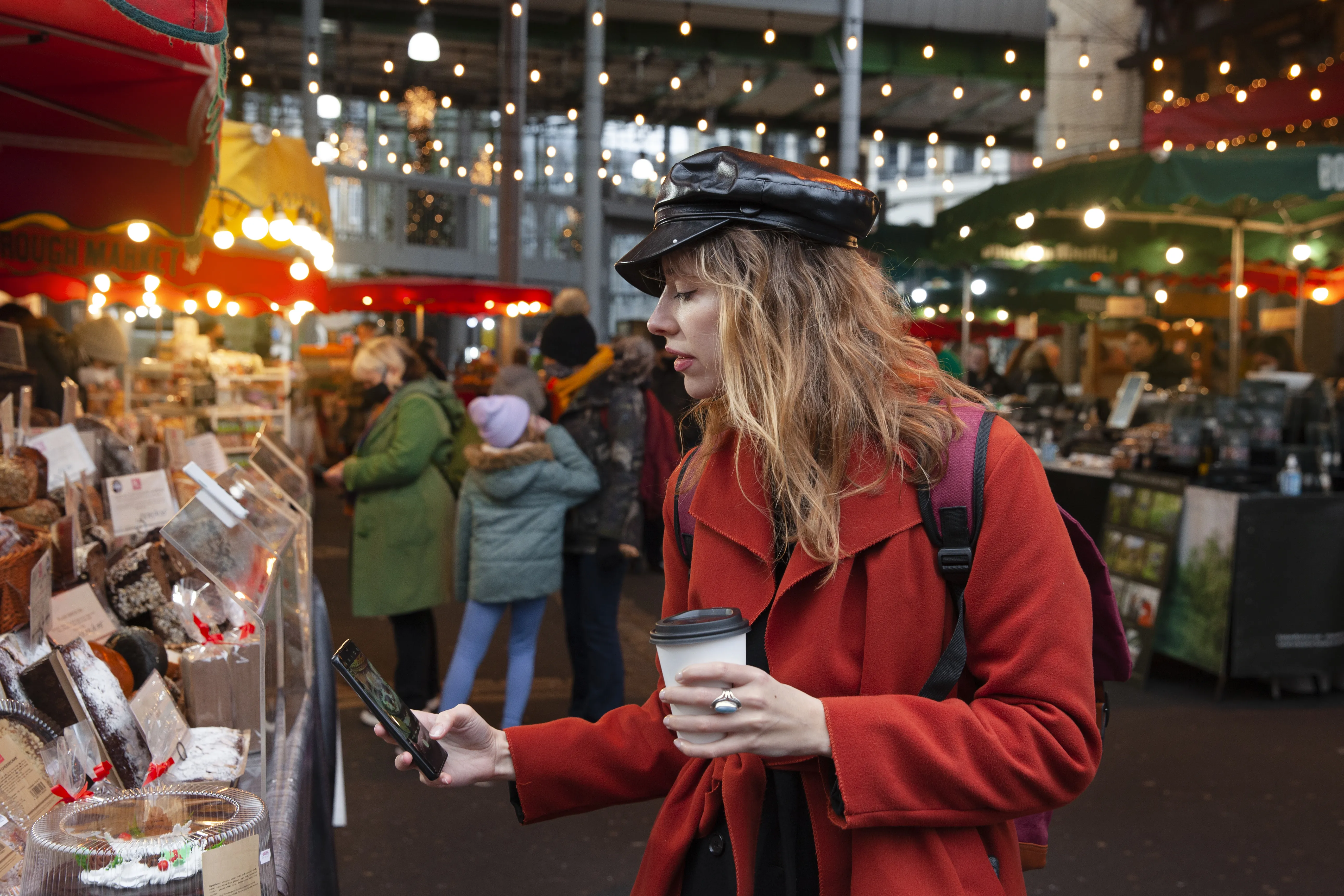 A medium profile shot of a woman with long wavy blonde hair wearing a black leather newsboy cap and a red coat, standing in an outdoor market.