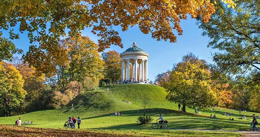 A wide-angle, eye-level shot of the Monopteros temple sitting atop a grassy hill in the English Garden (Englischer Garten) in Munich, Germany, during autumn.