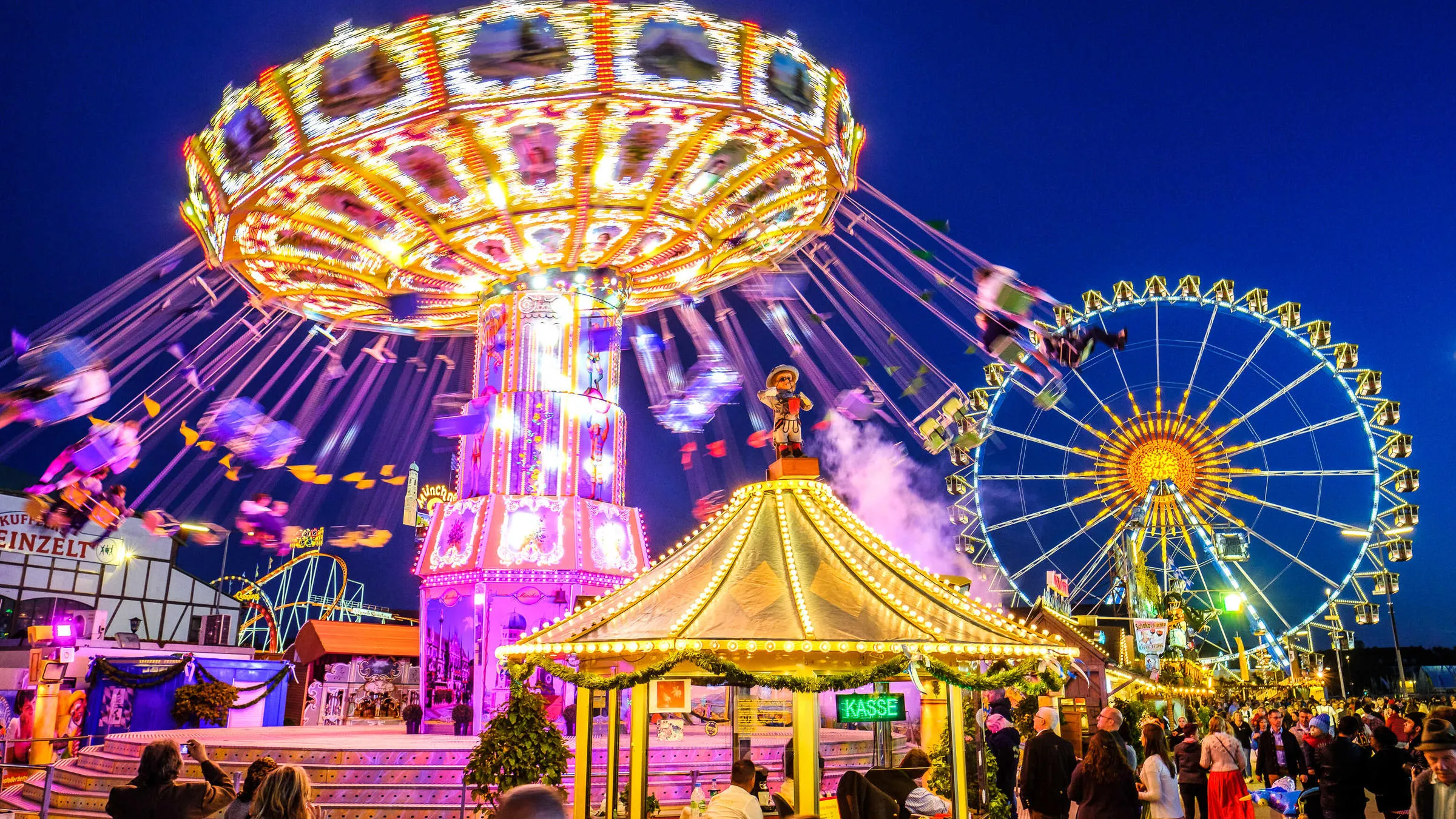 A low-angle, wide-angle long exposure shot of a vibrant amusement park at night, dominated by a spinning wave swinger ride on the left creating bright yellow and pink light trails.