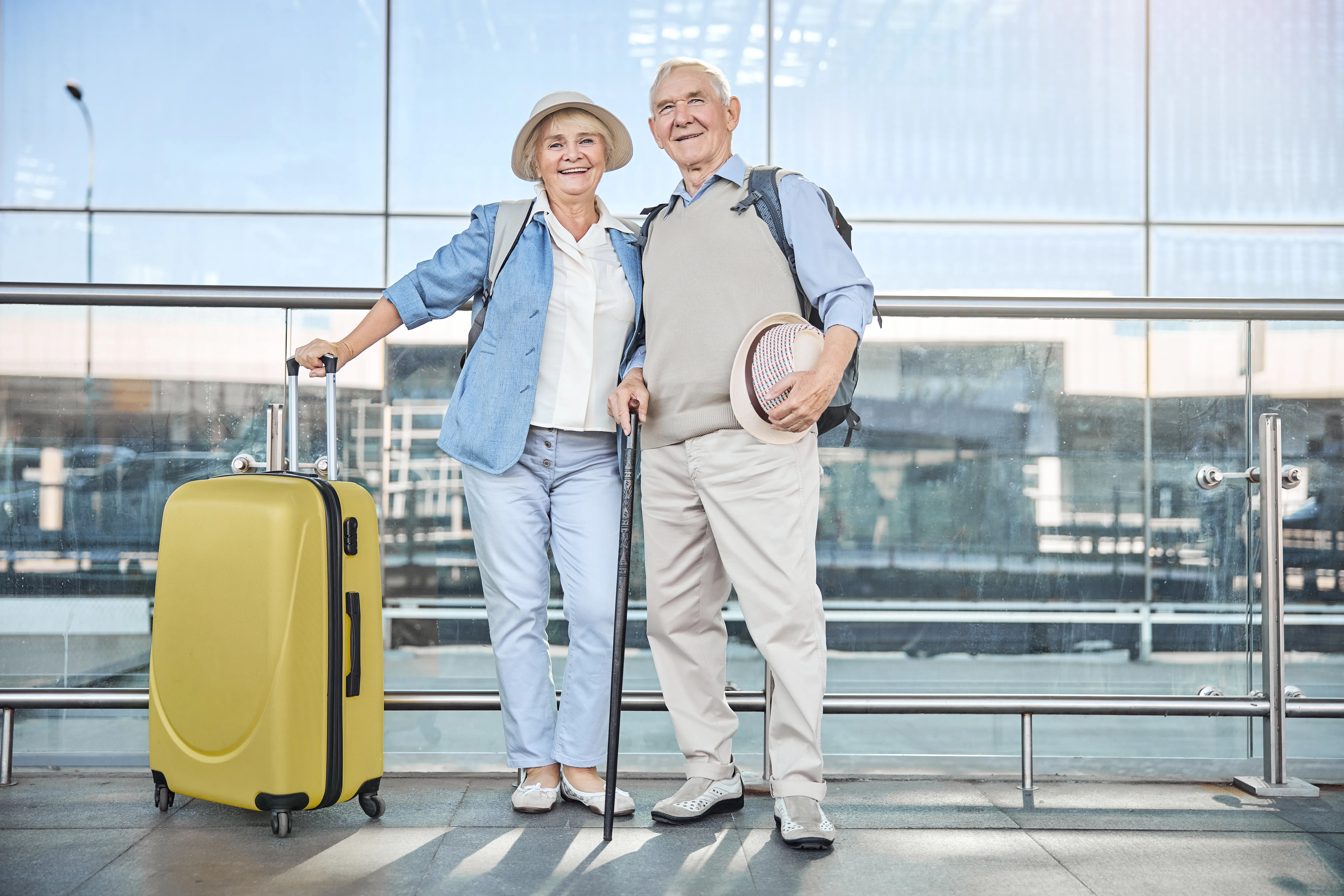 Two senior travelers standing at an airport with a yellow suitcase and a cane