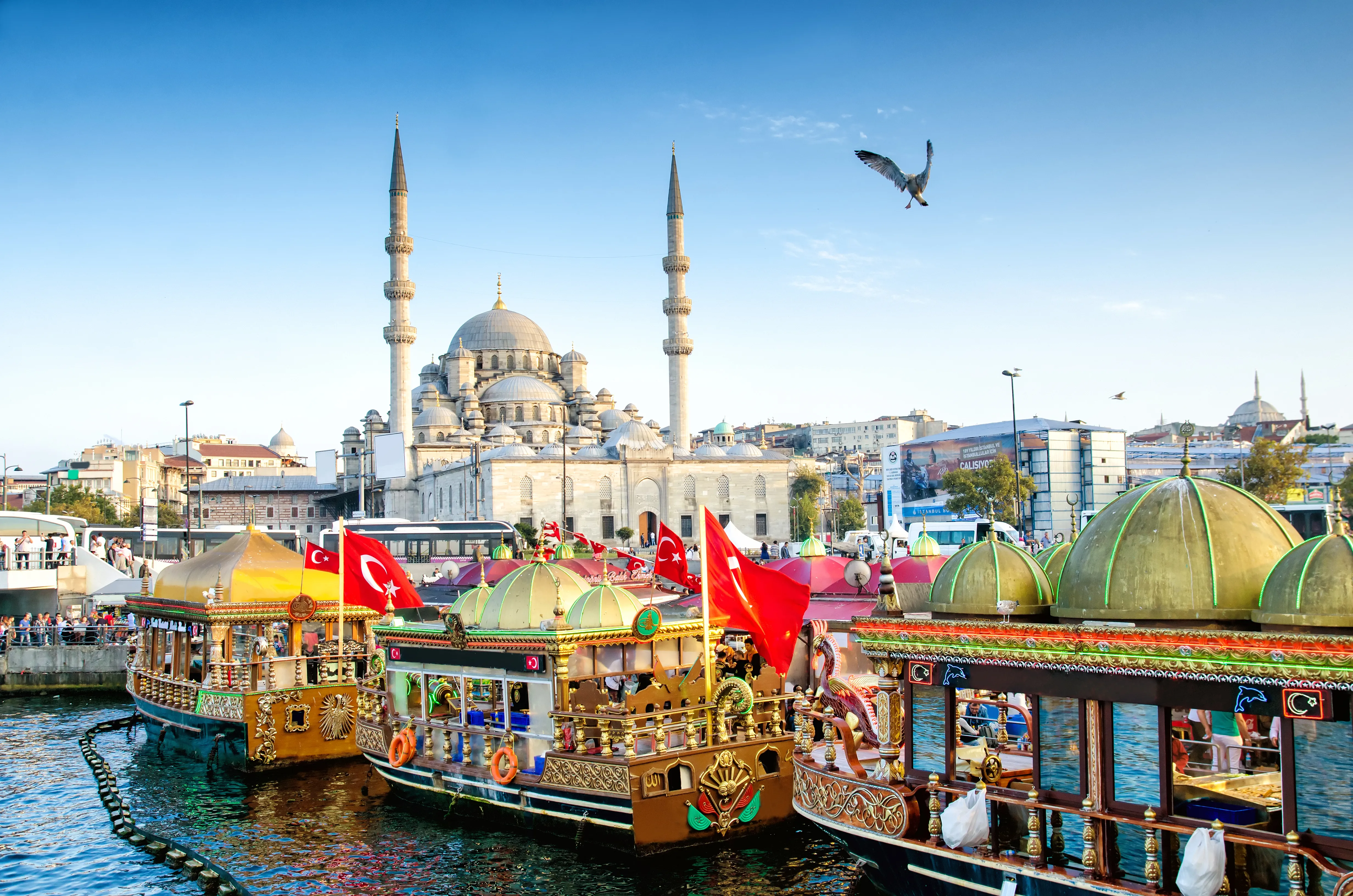 An architectural and cultural photograph shows the New Mosque (Yeni Cami) rising behind traditional floating fish boats at the Eminönü waterfront in Istanbul, Turkey.