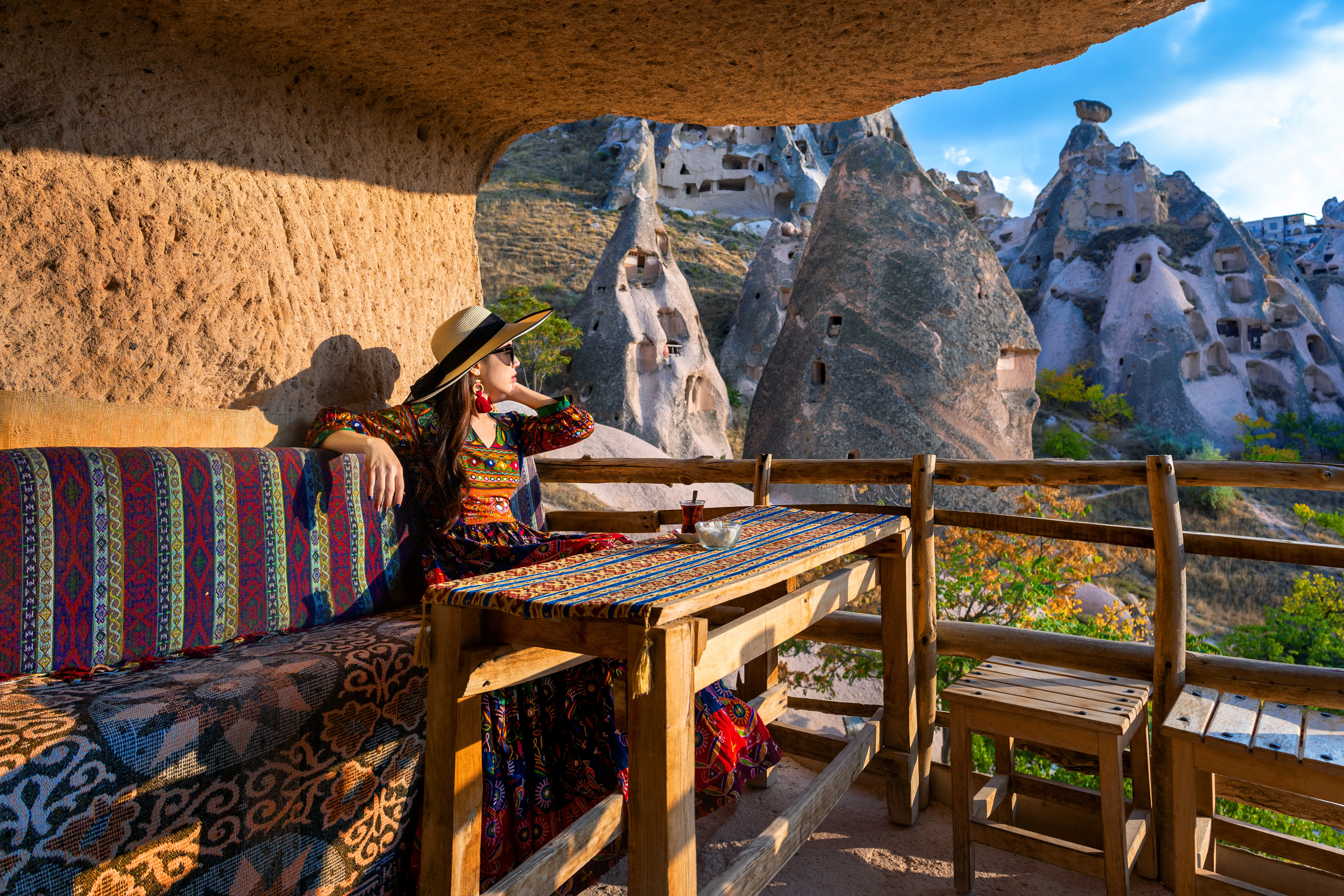  A travel and lifestyle photograph shows a woman relaxing at an outdoor cave terrace overlooking the historic volcanic rock formations of Cappadocia, Turkey.