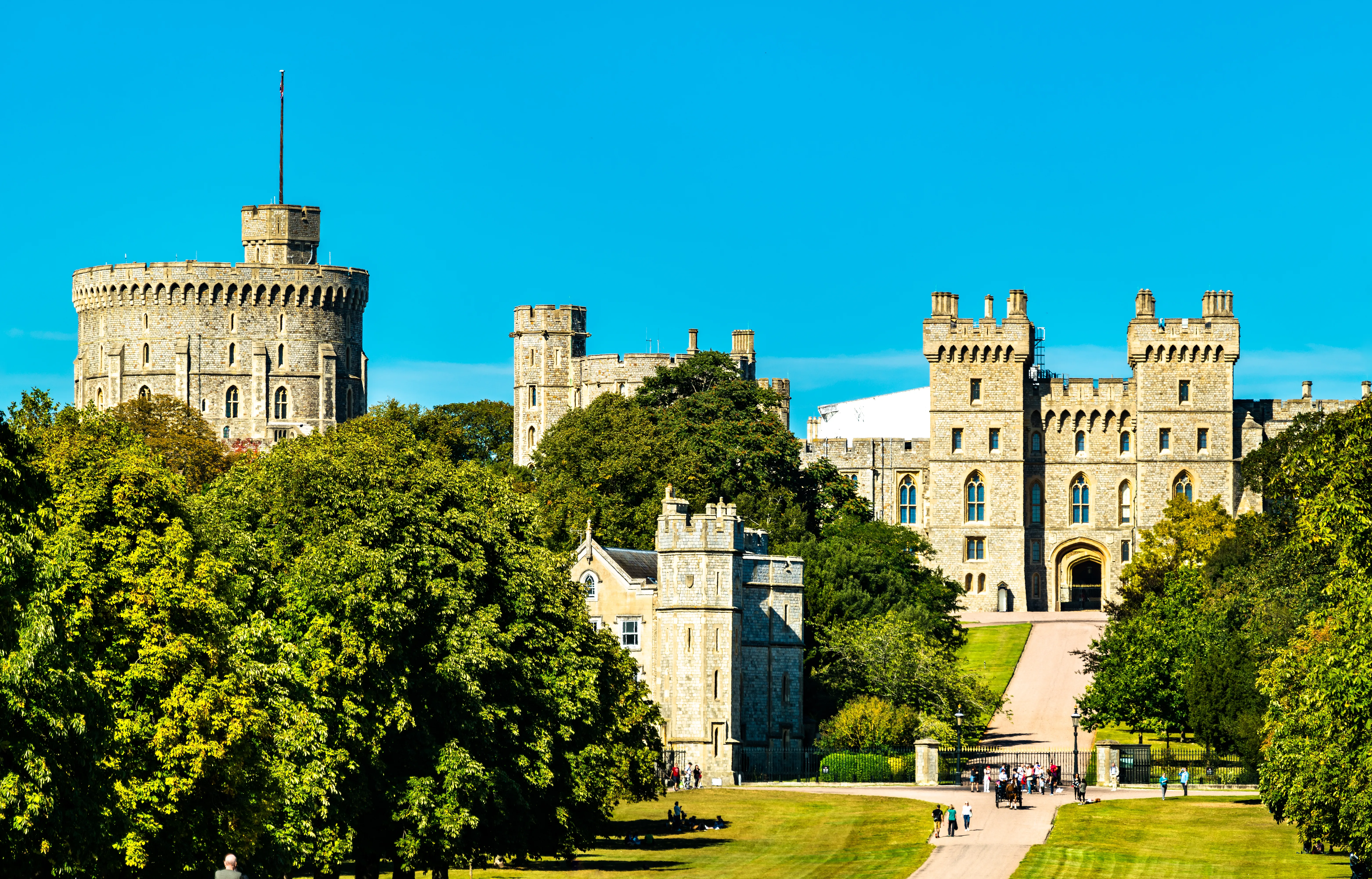 An architectural and landscape photograph shows Windsor Castle rising behind lush green trees and lawns in Windsor, Berkshire, United Kingdom