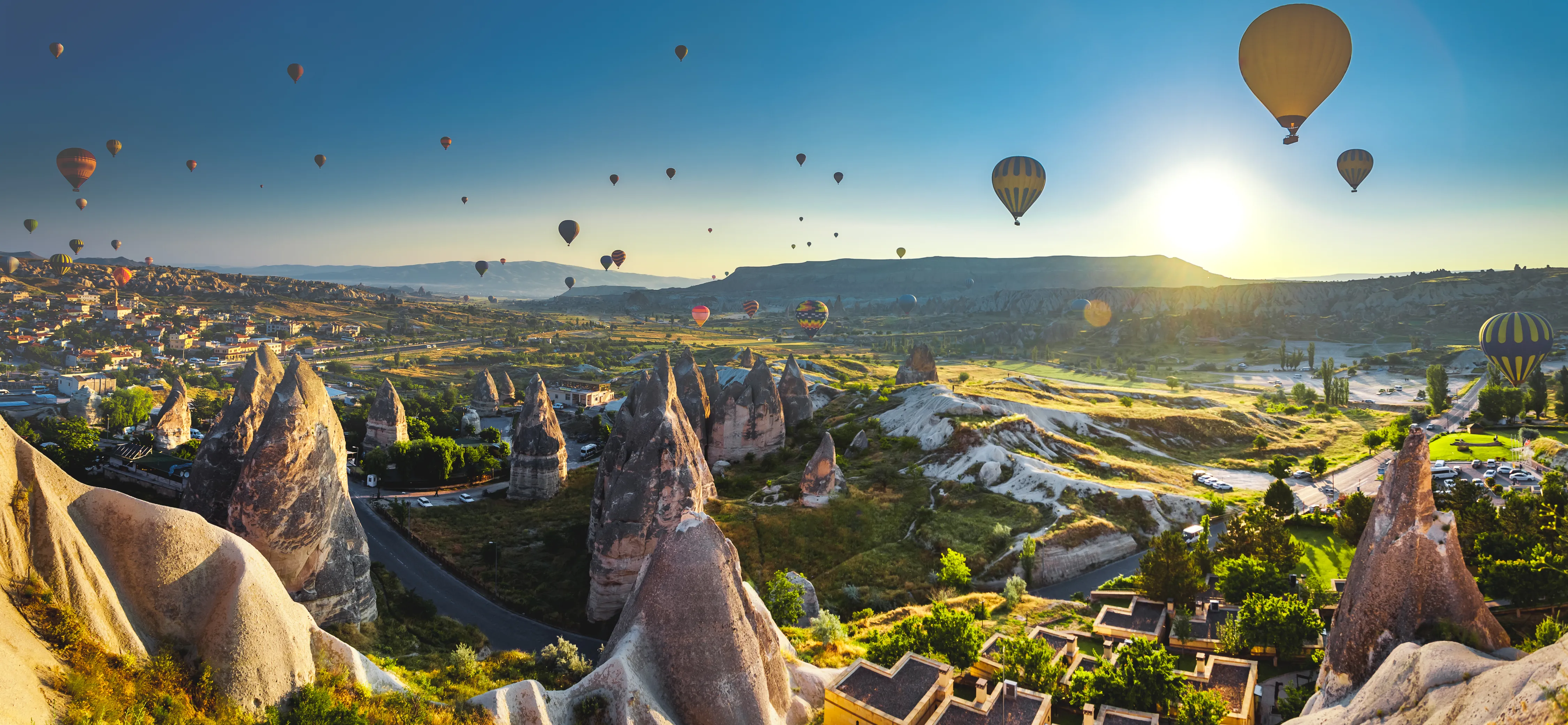 A landscape and travel photograph shows dozens of hot air balloons floating over the volcanic rock valleys of Cappadocia, Turkey, at sunrise