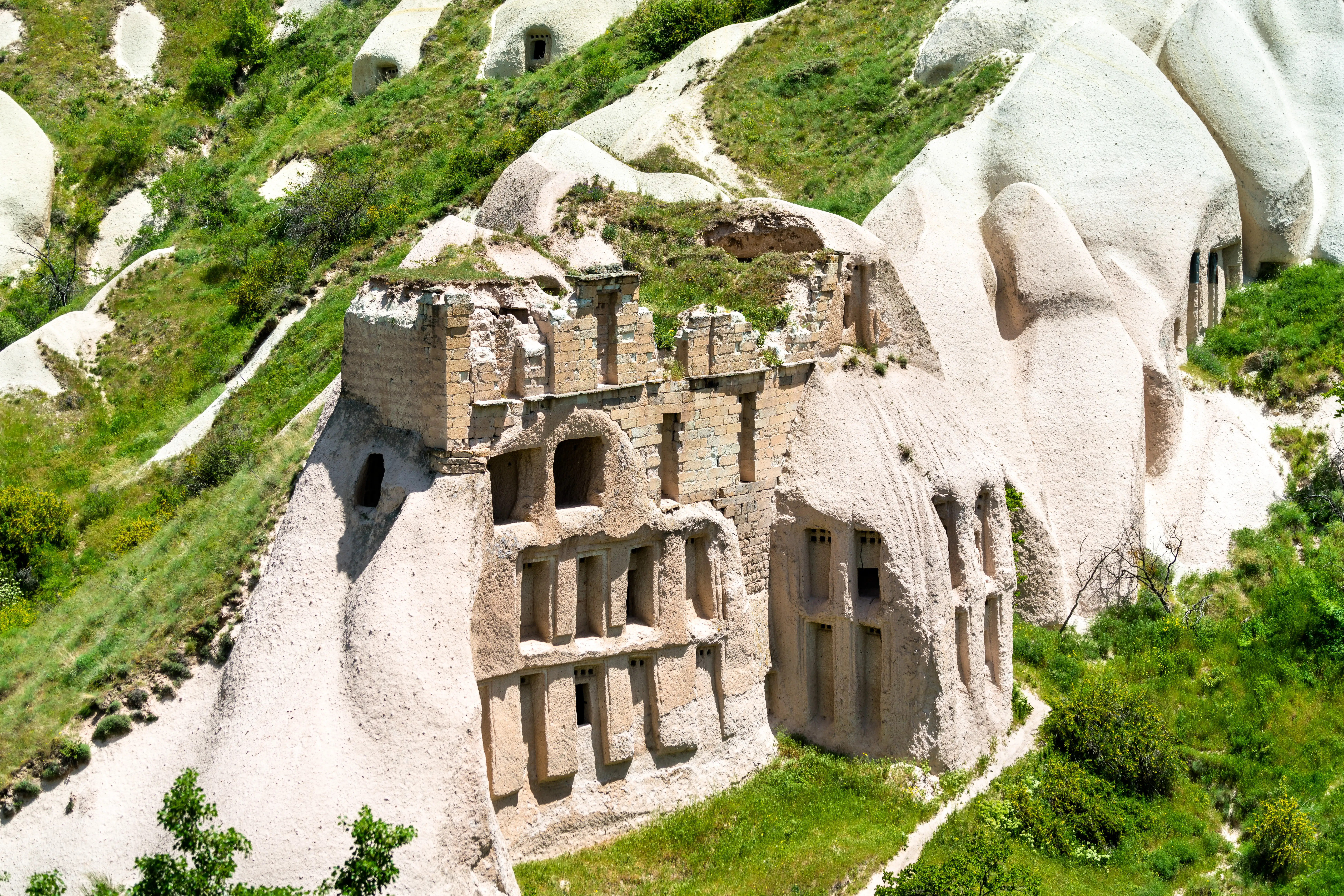 A high-angle architectural and landscape photograph shows an ancient, crumbling stone and rock-cut structure built into the volcanic formations of Cappadocia, Turkey