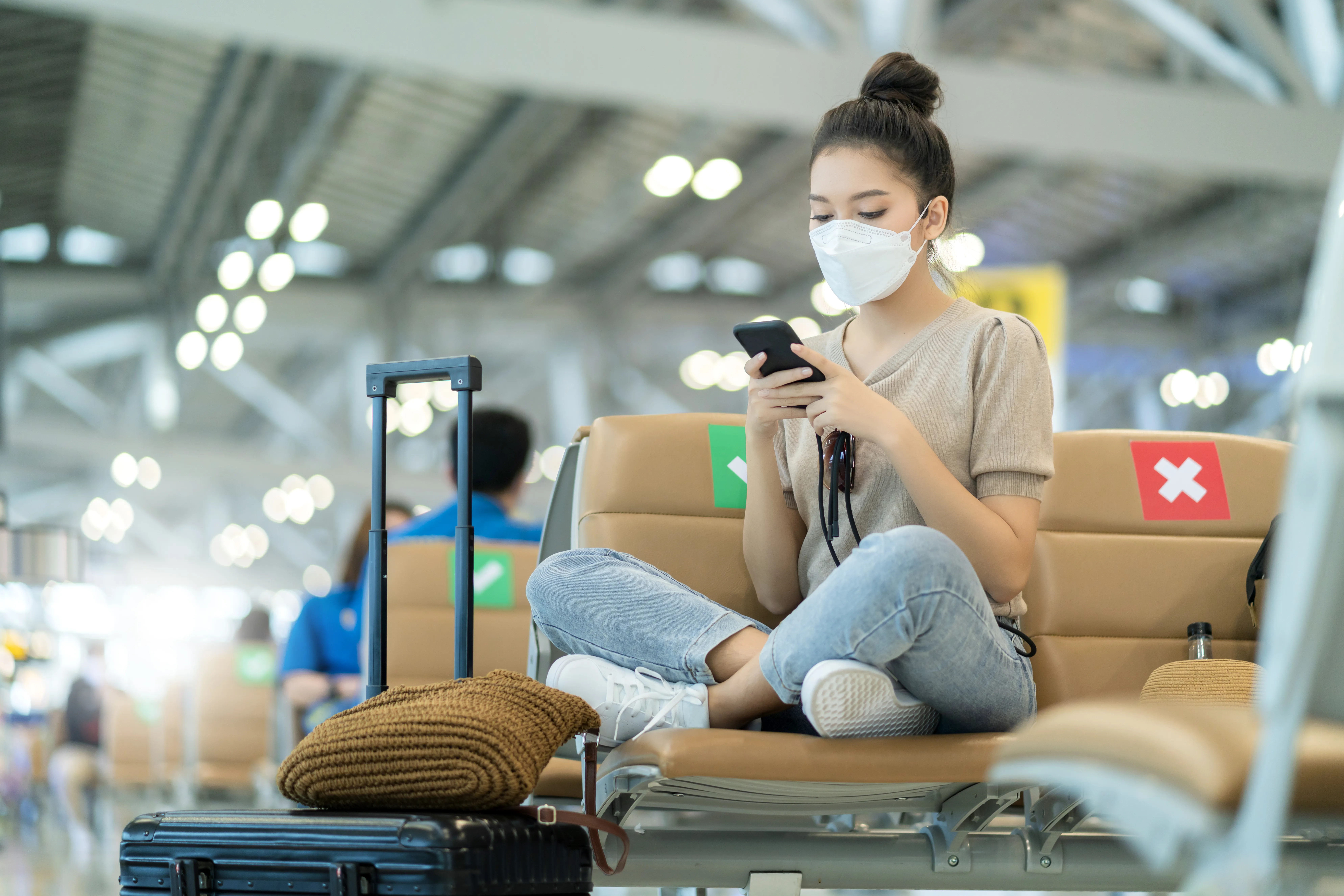 A masked young woman sits cross-legged at an airport terminal bench, looking at her smartphone next to her luggage
