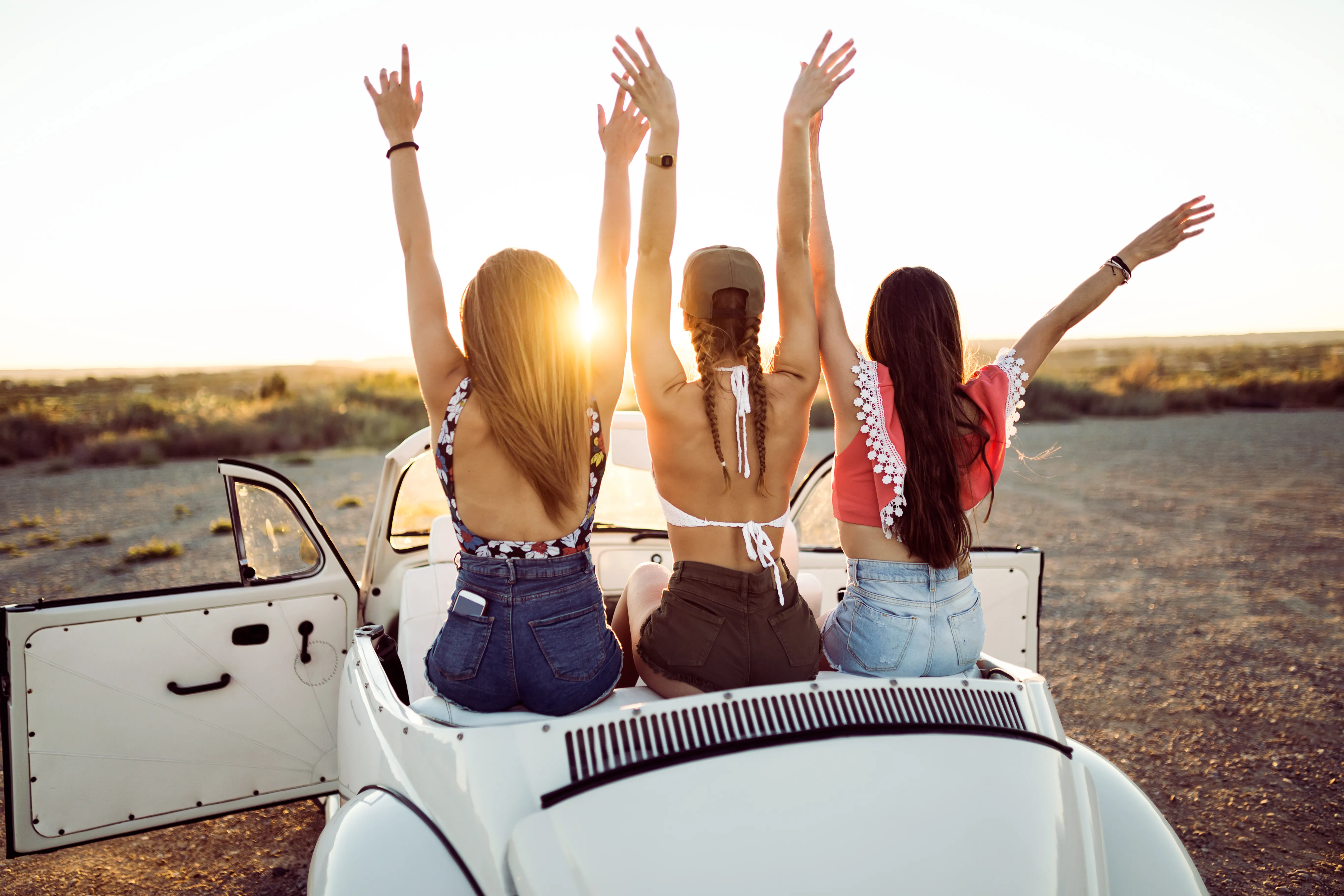 Three young women sitting on the back of a white convertible car with their arms raised toward the sunset