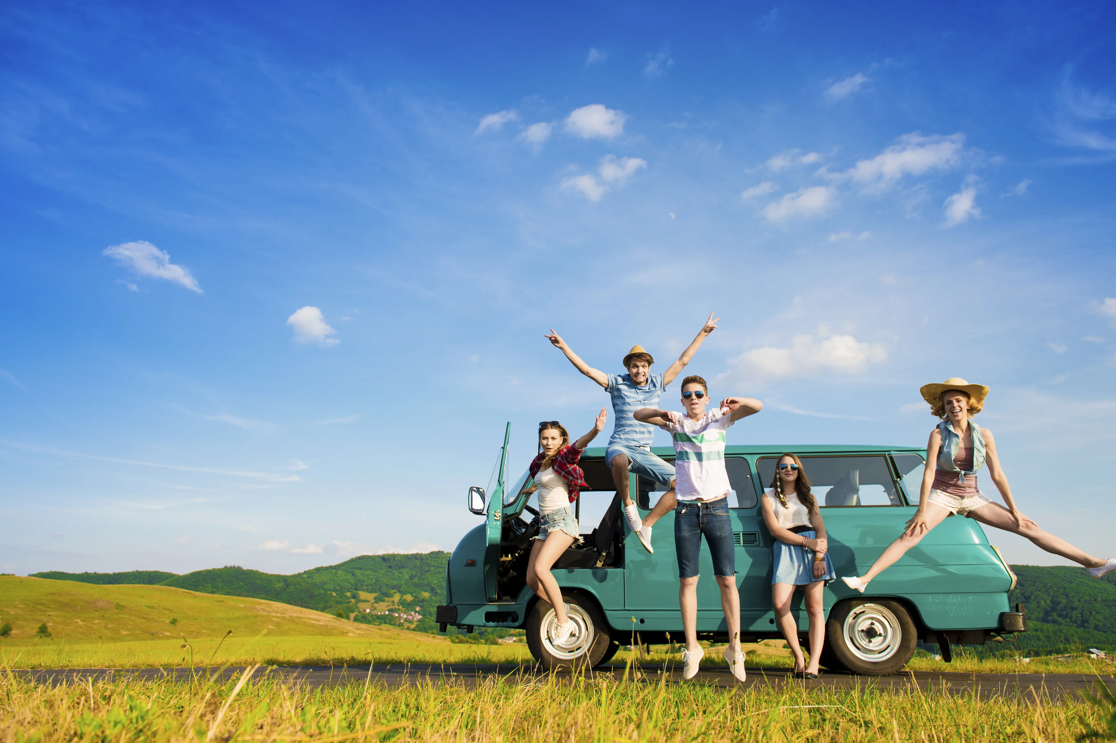 A group of joyful young friends jumping and posing around a teal van on a scenic road trip