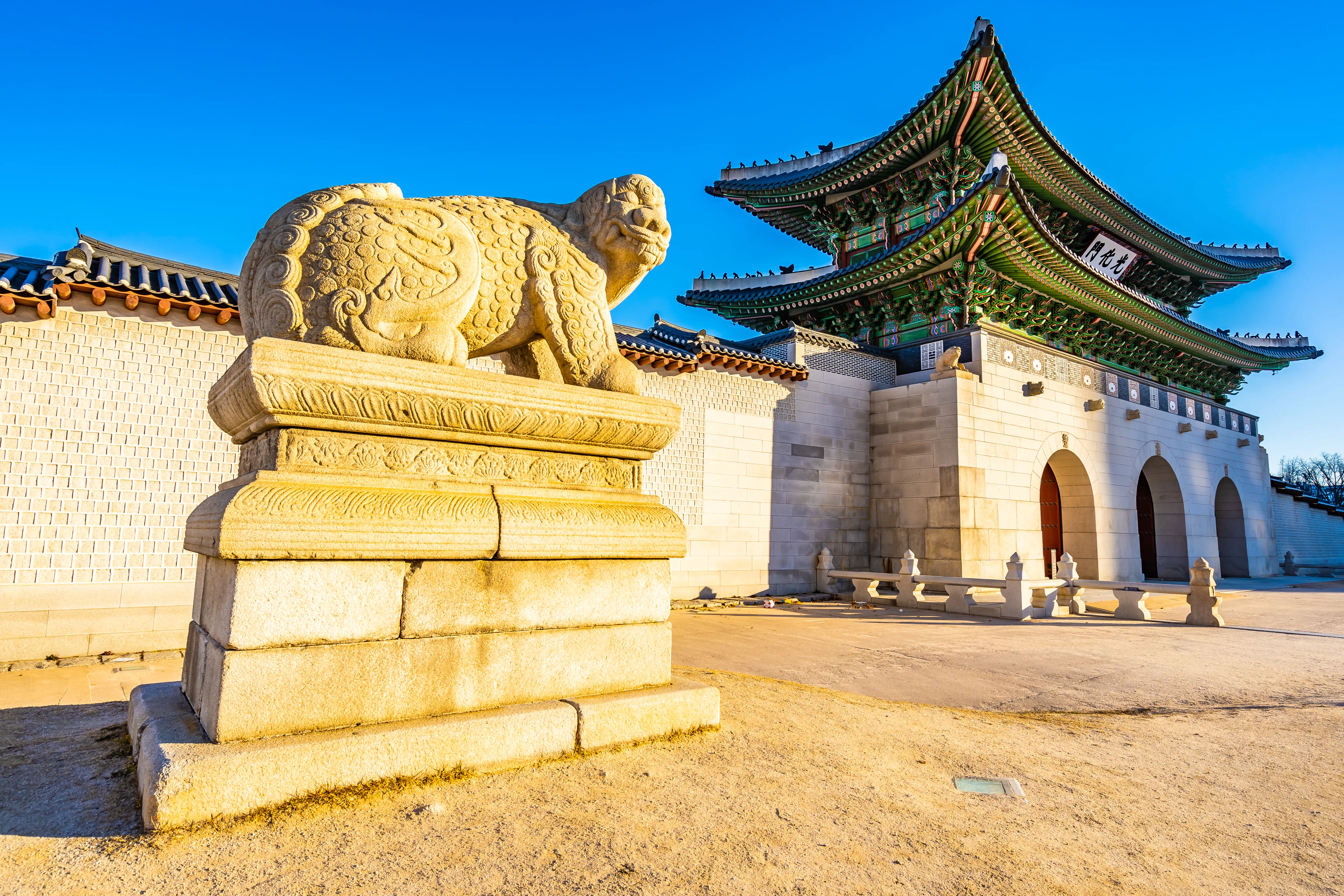 Stone guardian lion in front of Gyeongbokgung Palace's main gate in Seoul, South Korea. Clear blue sky emphasizes the historic architecture.