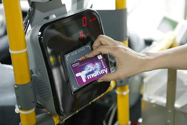 A hand holds a purple T-money card, tapping it on a card reader inside a vehicle. The setting suggests public transport, with yellow poles visible.
