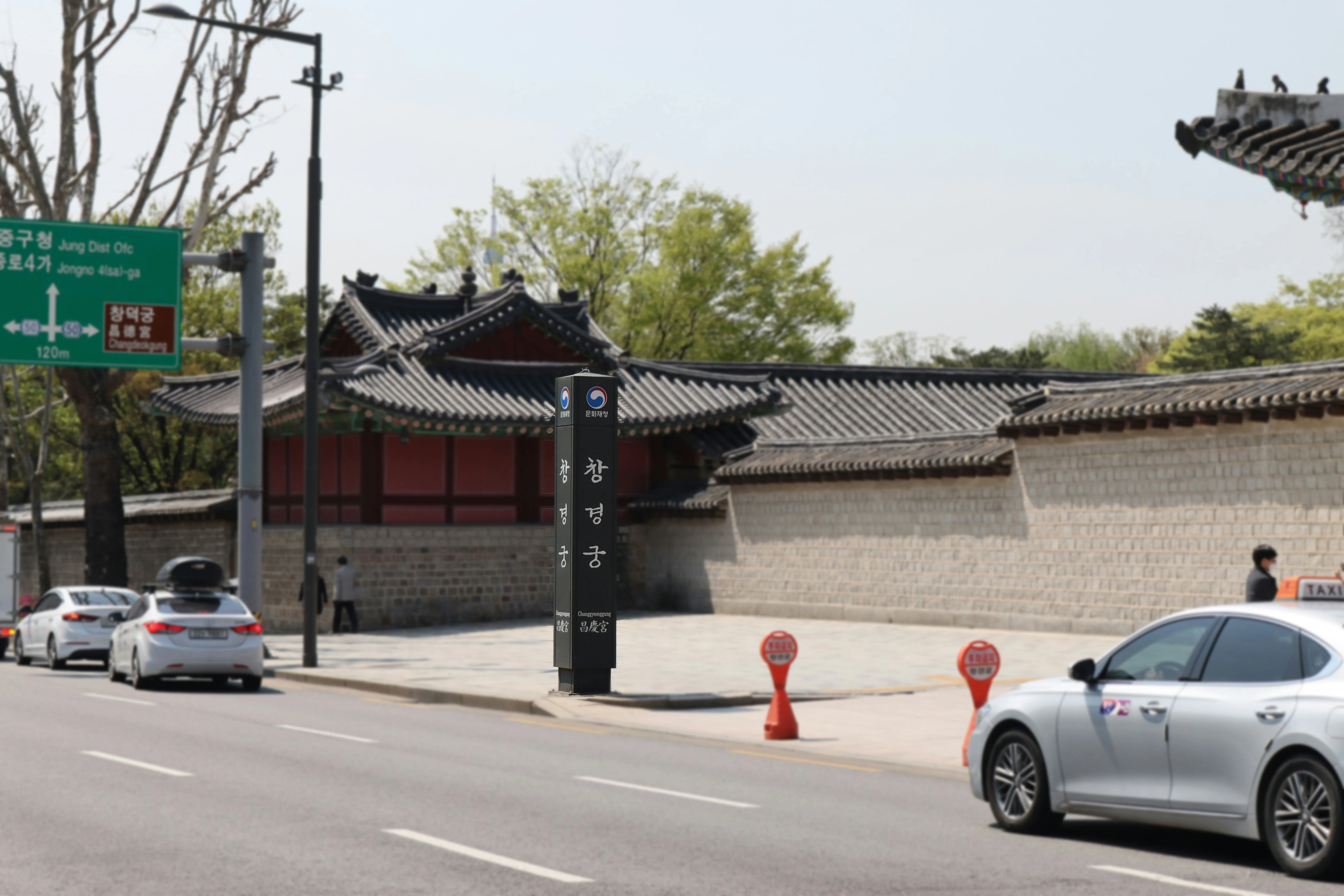 Street outside traditional Korean palace, lined with a stone wall and ornate roofs. Cars and people pass by on a sunny day. Signs are visible in Korean