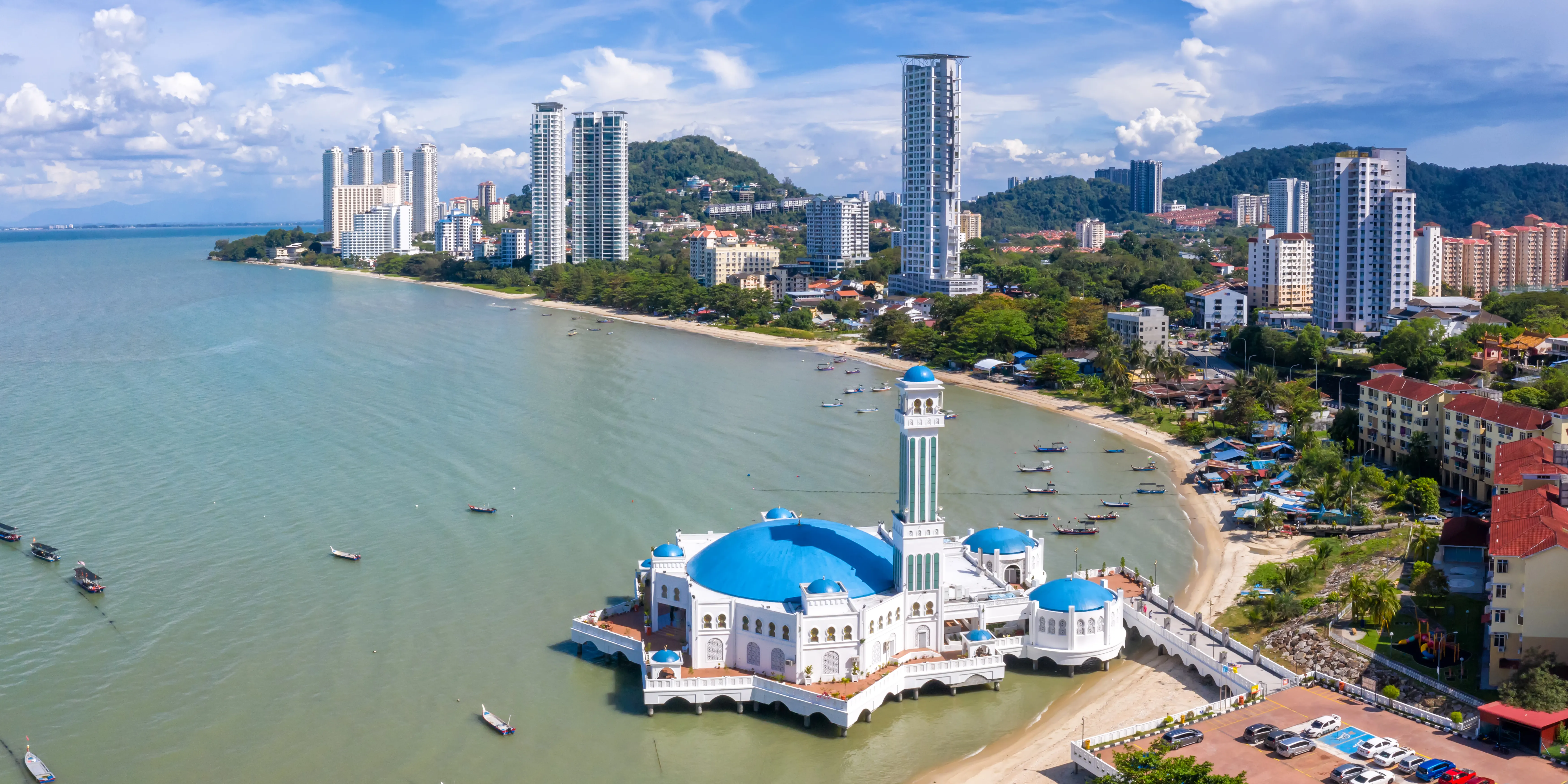 Aerial view of a white floating mosque with blue domes on a coastal shoreline next to a city. 