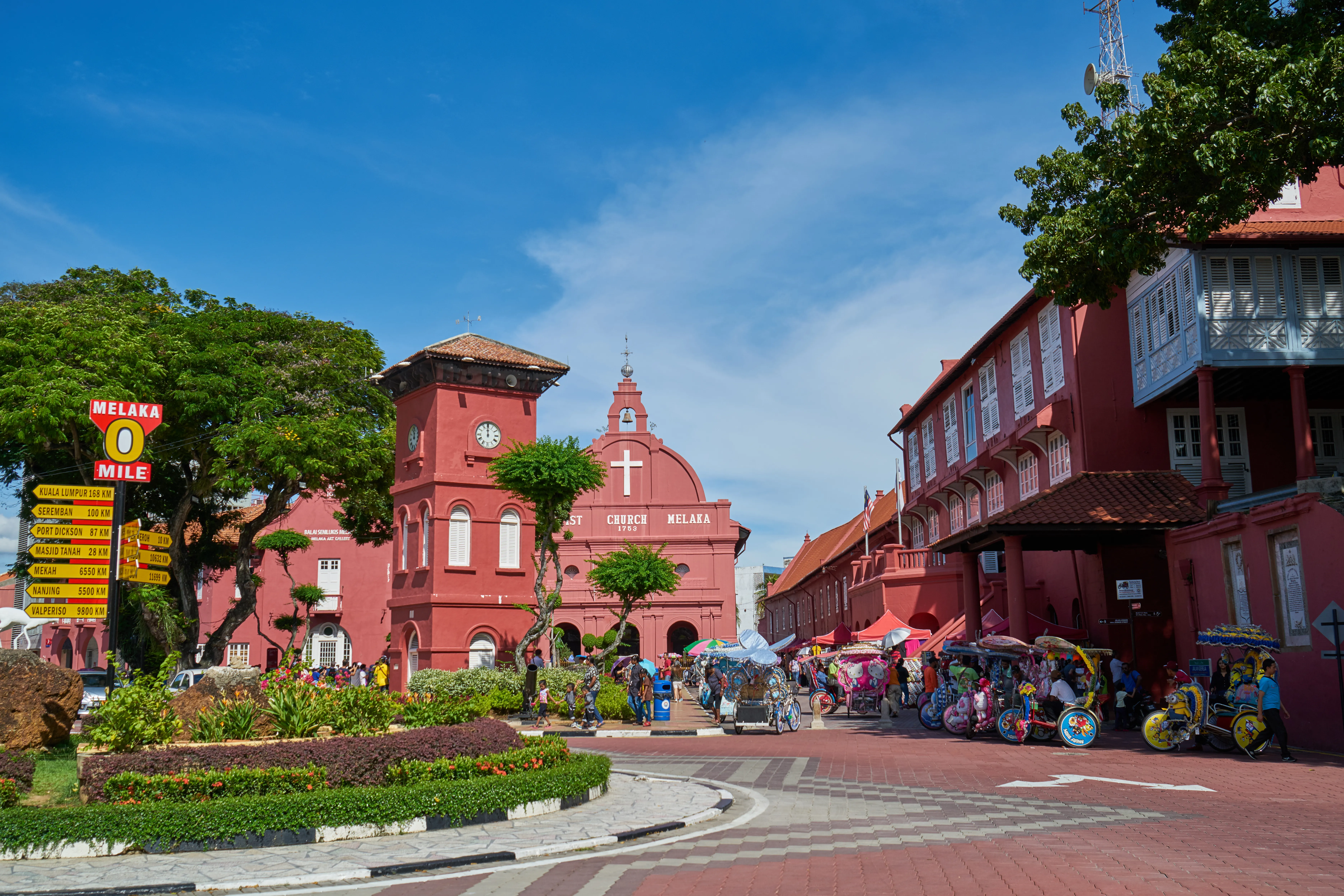 A historic red church and clock tower in a bustling square with decorated trishaws under a blue sky