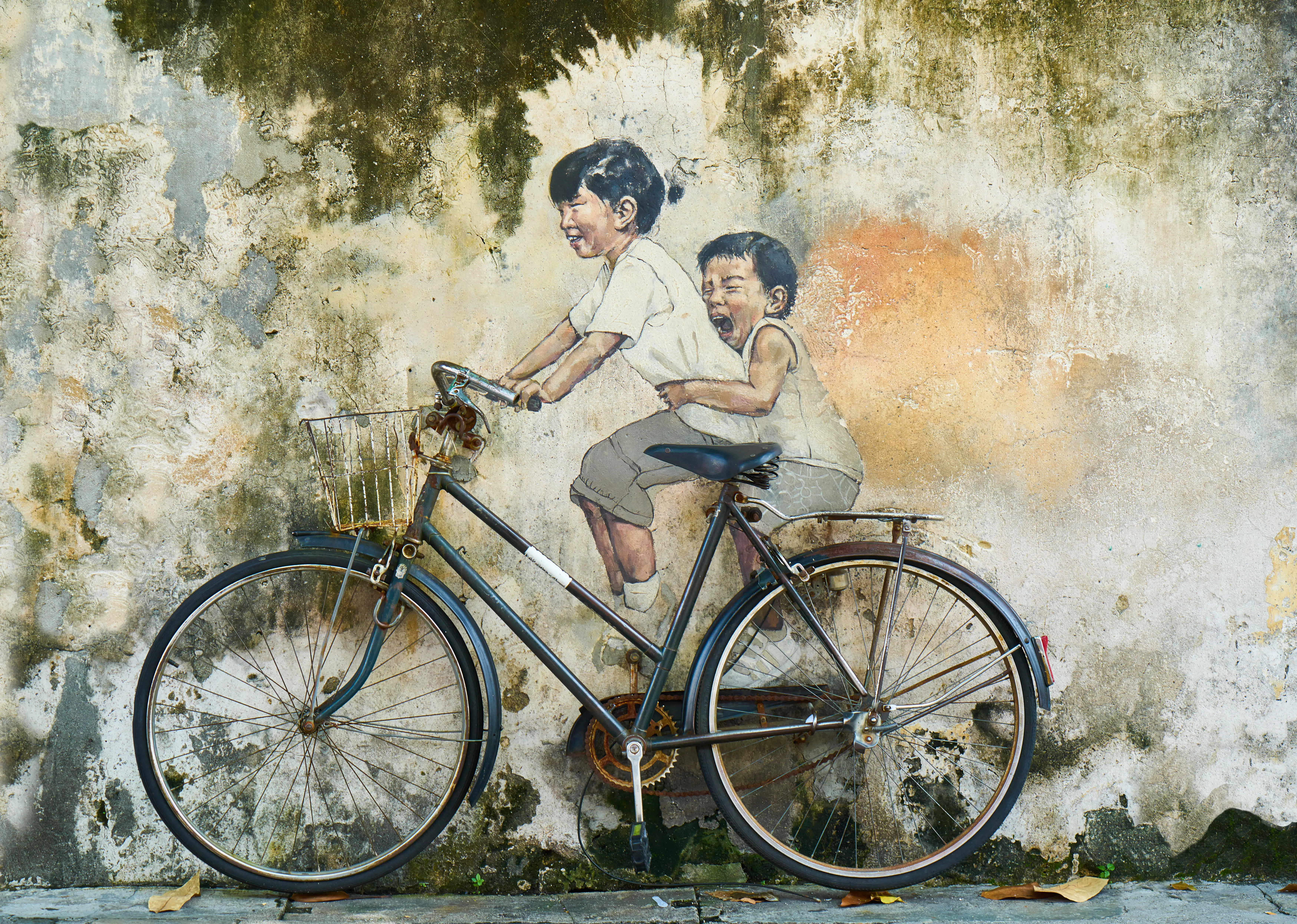 Street art mural of two children painted on a weathered wall, interacting with a real bicycle