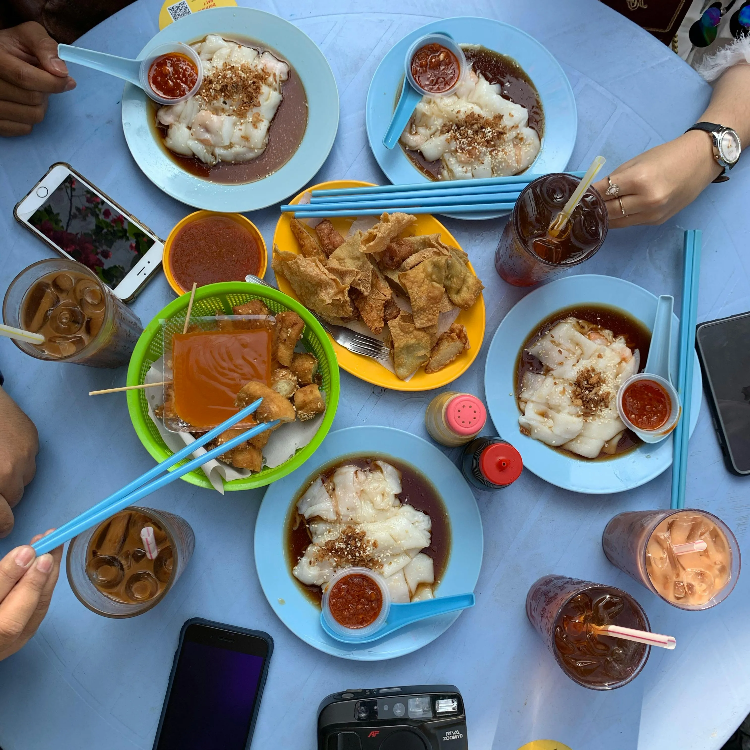 Overhead view of a blue table with Malaysian Chee Cheong Fun, fried snacks, iced drinks, and hands reaching with chopsticks