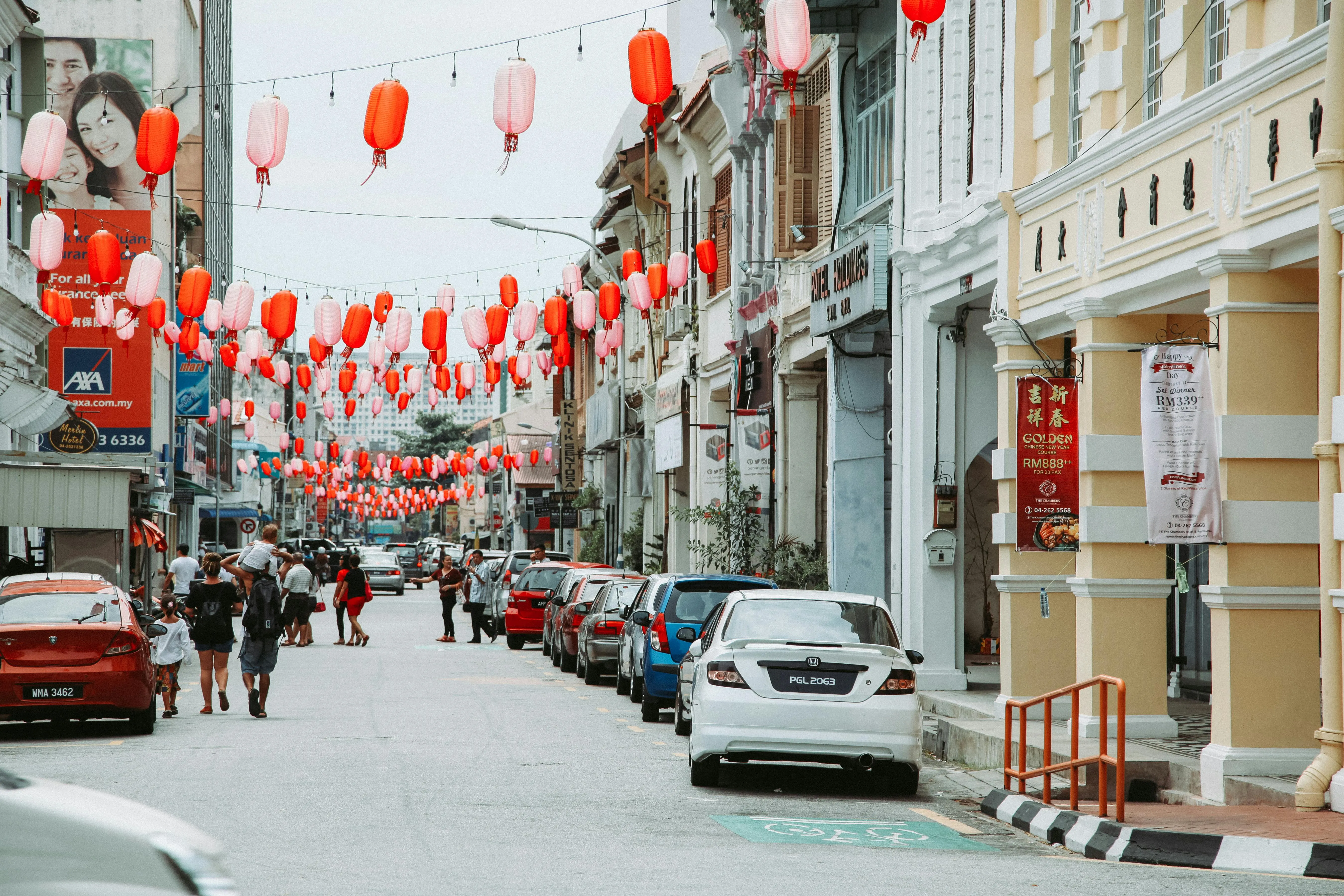 A vibrant city street lined with historic shophouses, parked cars, and rows of red lanterns hanging overhead.