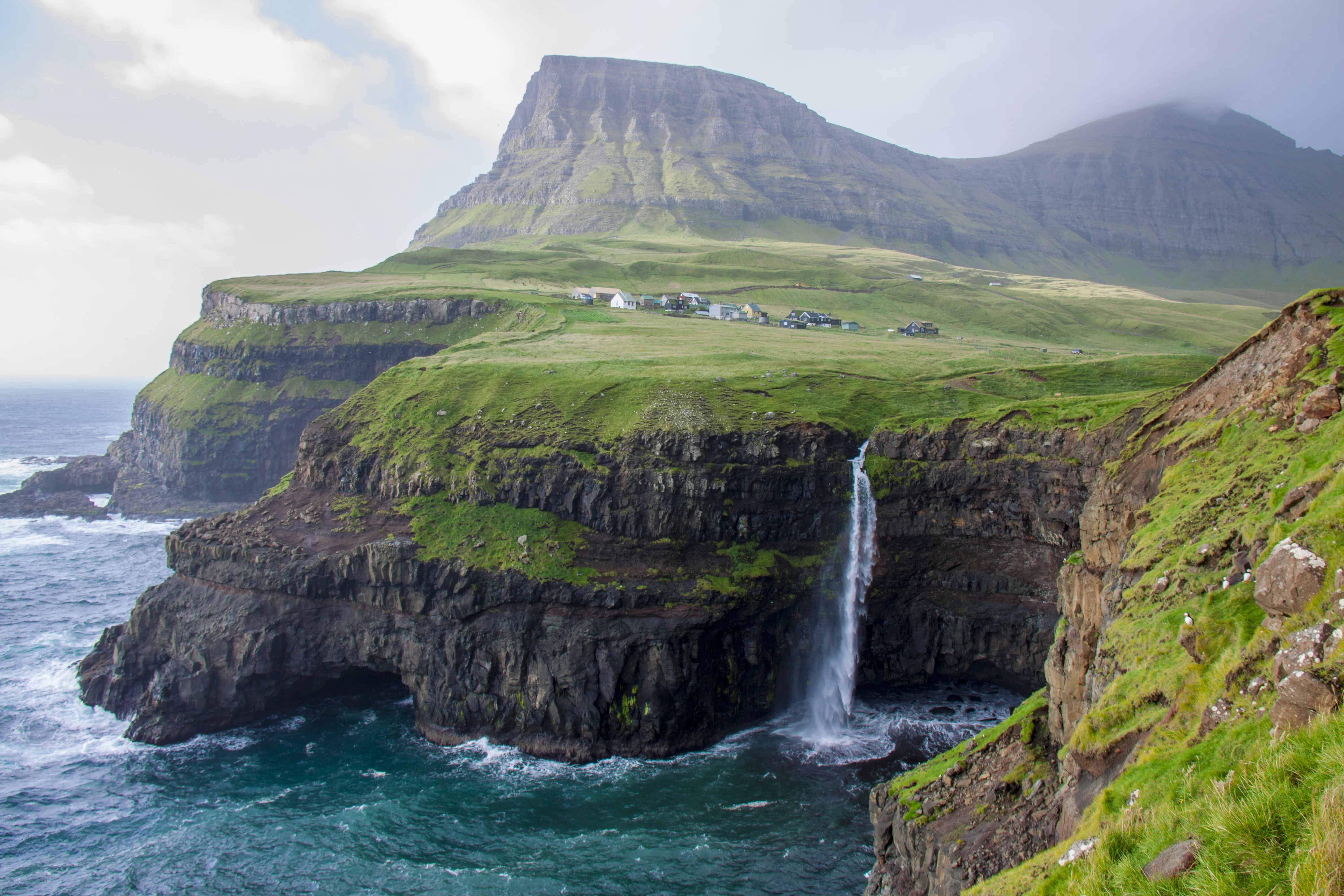 A serene landscape of a coastal cliff with a waterfall flowing into the ocean below. Above, grassy fields with a few houses sit under a cloudy sky.