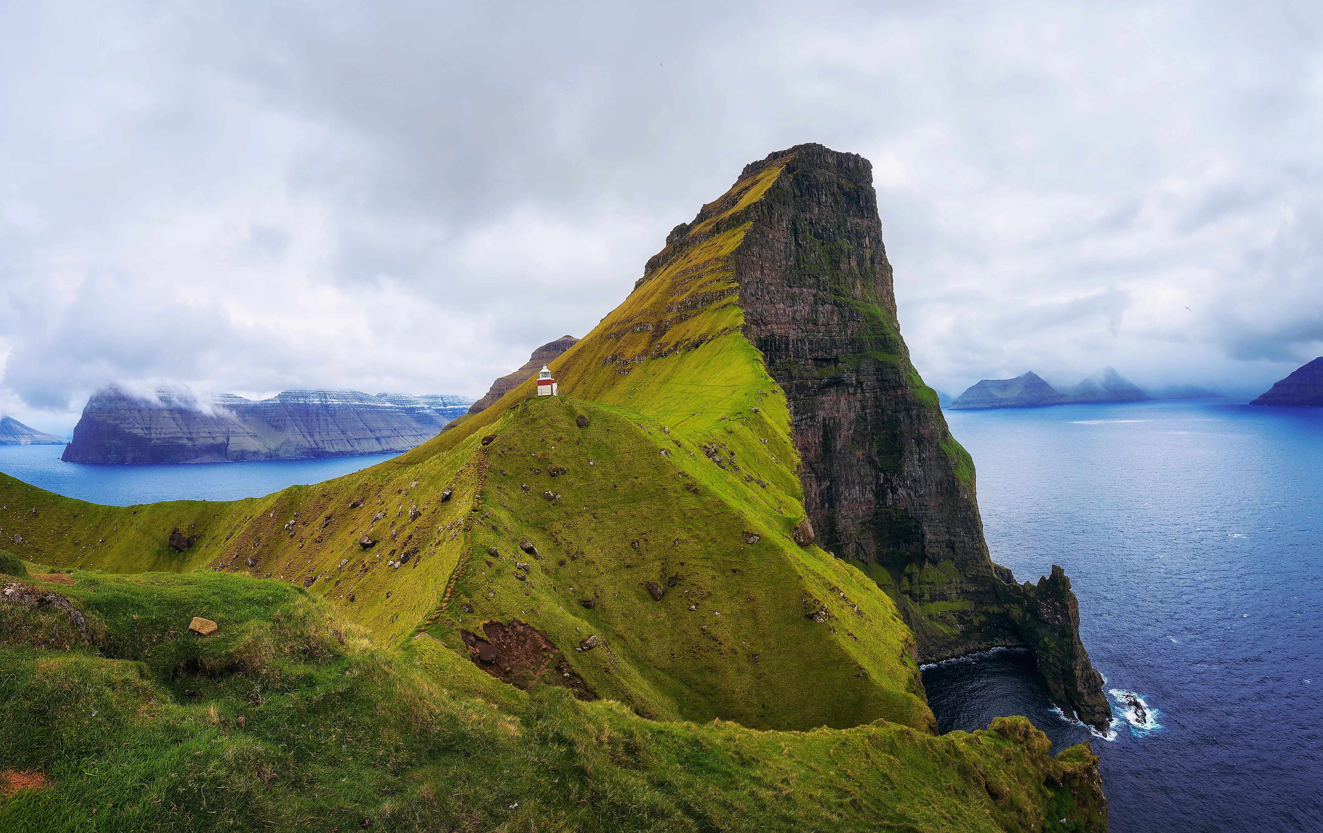 A lone lighthouse sits atop a green, grassy hill, leading to a steep cliff with the ocean below. Cloudy skies loom over distant misty islands, creating a serene, remote atmosphere