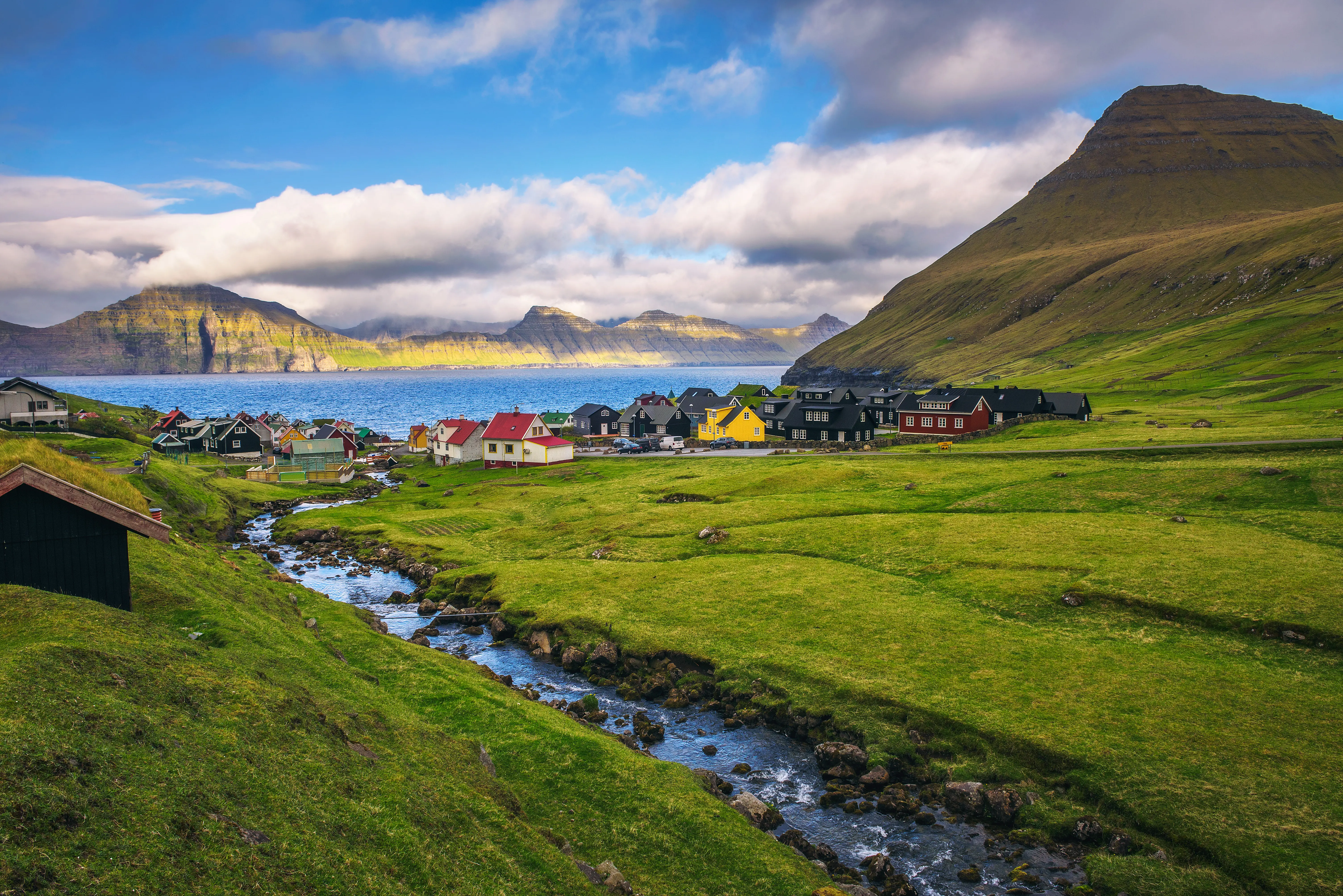 A picturesque village with colorful houses sits by a sparkling stream, surrounded by lush green hills and a distant mountain under a blue sky