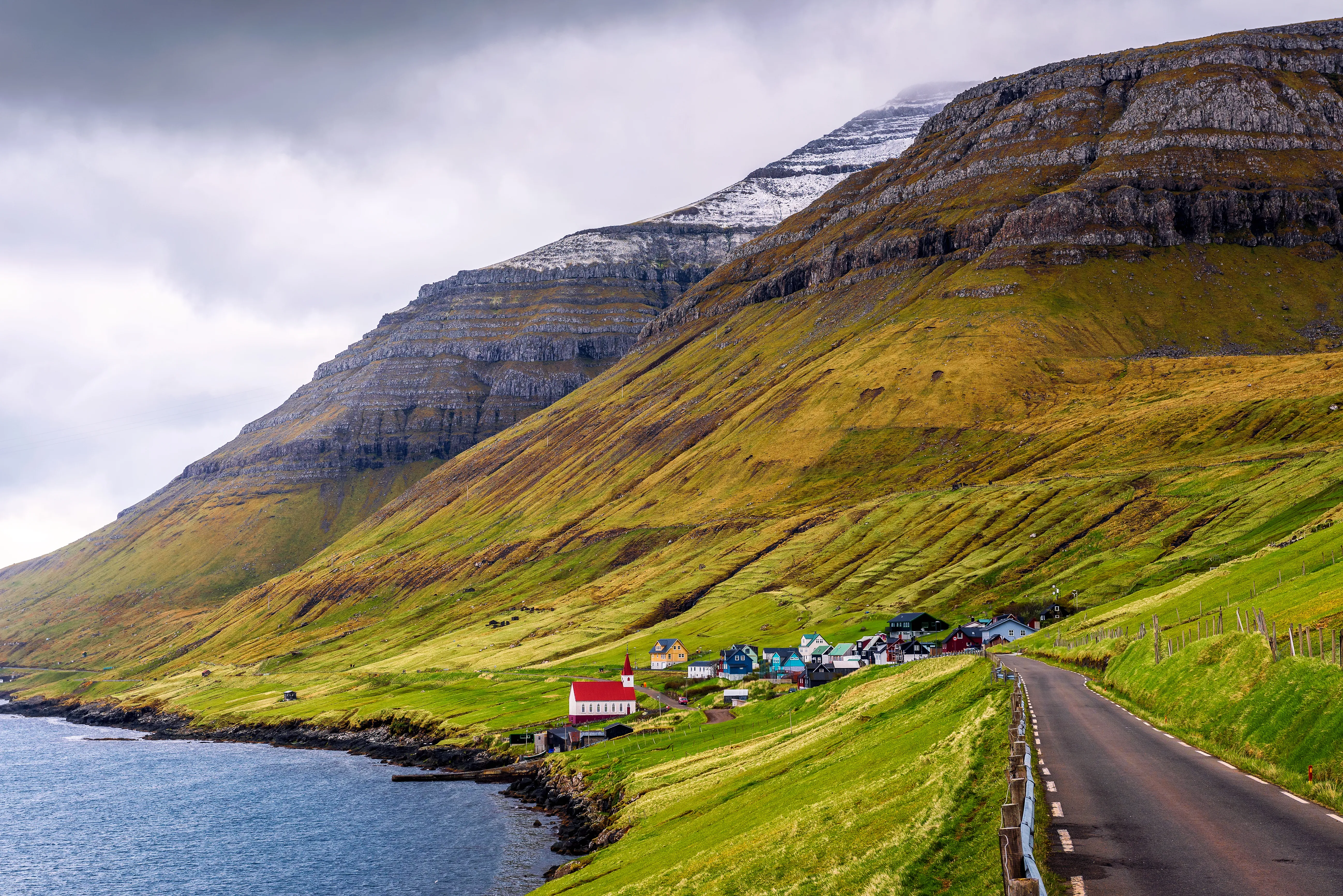 Coastal village with colorful houses nestled under steep, grassy hills. A winding road and overcast sky add a serene and remote feel to the scene