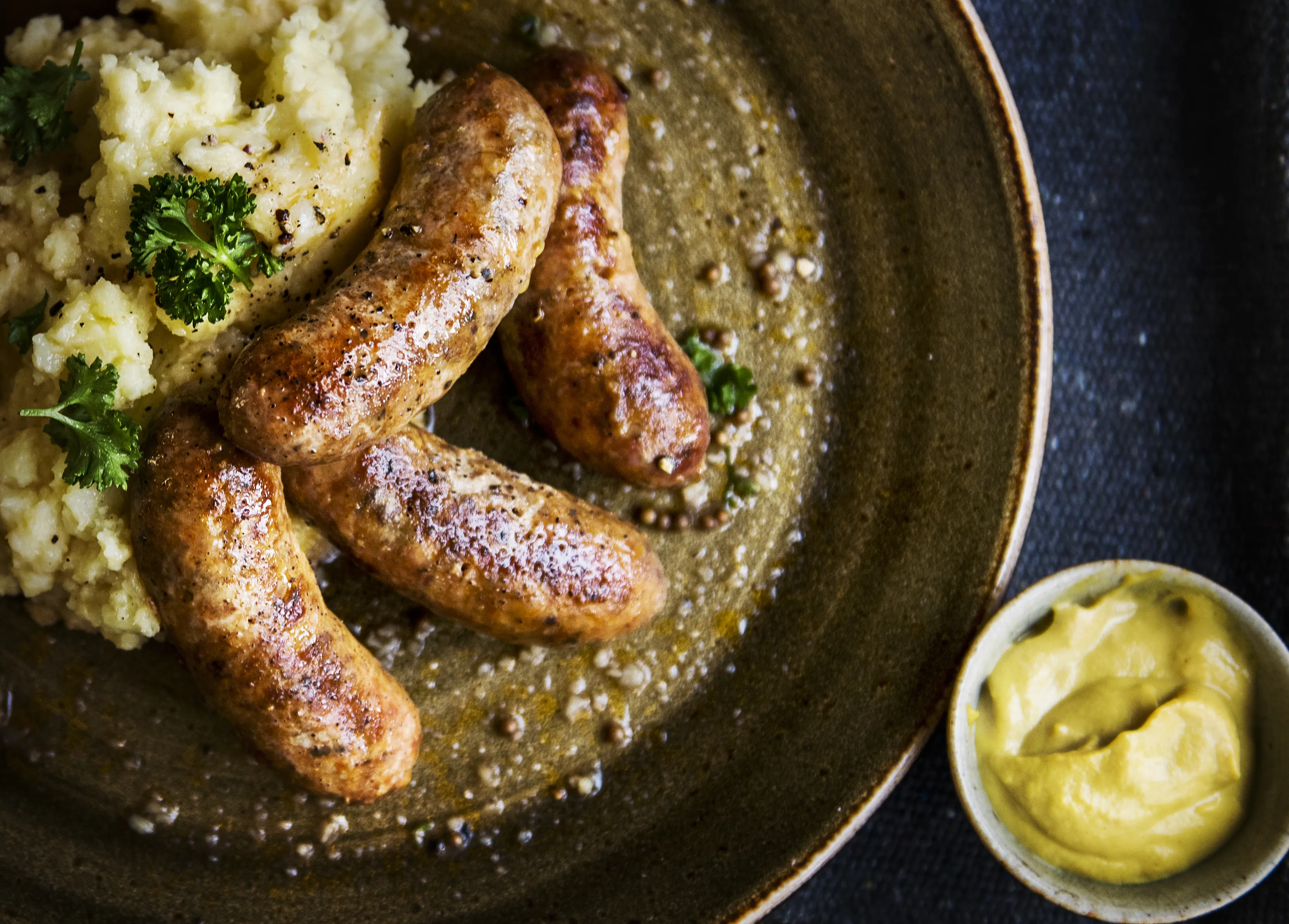Plate of golden-brown sausages and creamy mashed potatoes garnished with parsley, accompanied by a small bowl of mustard. Cozy, hearty meal