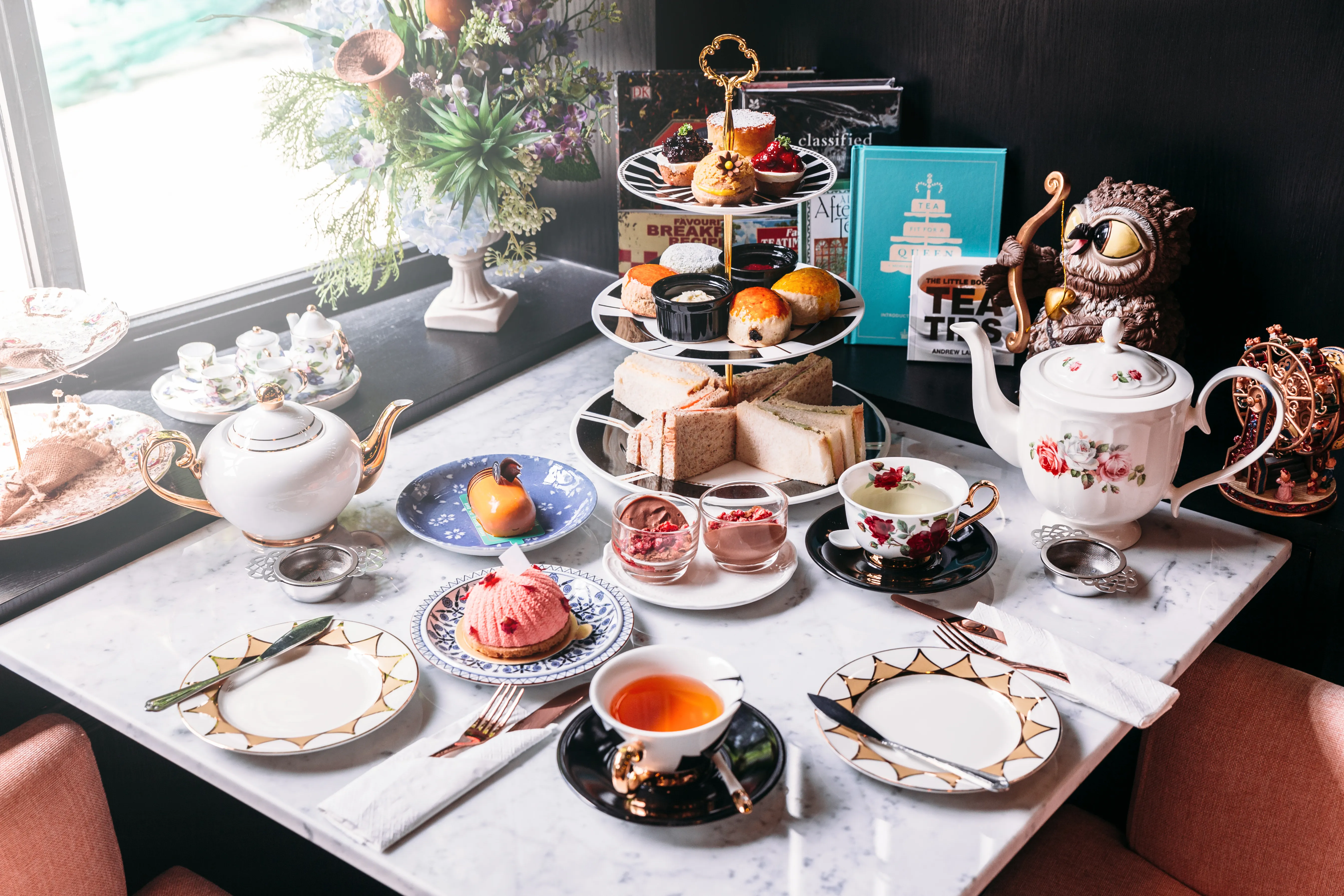 A beautifully set afternoon tea scene with a tiered tray of pastries, sandwiches, and scones on a marble table. Elegant teapots and floral teacups add charm