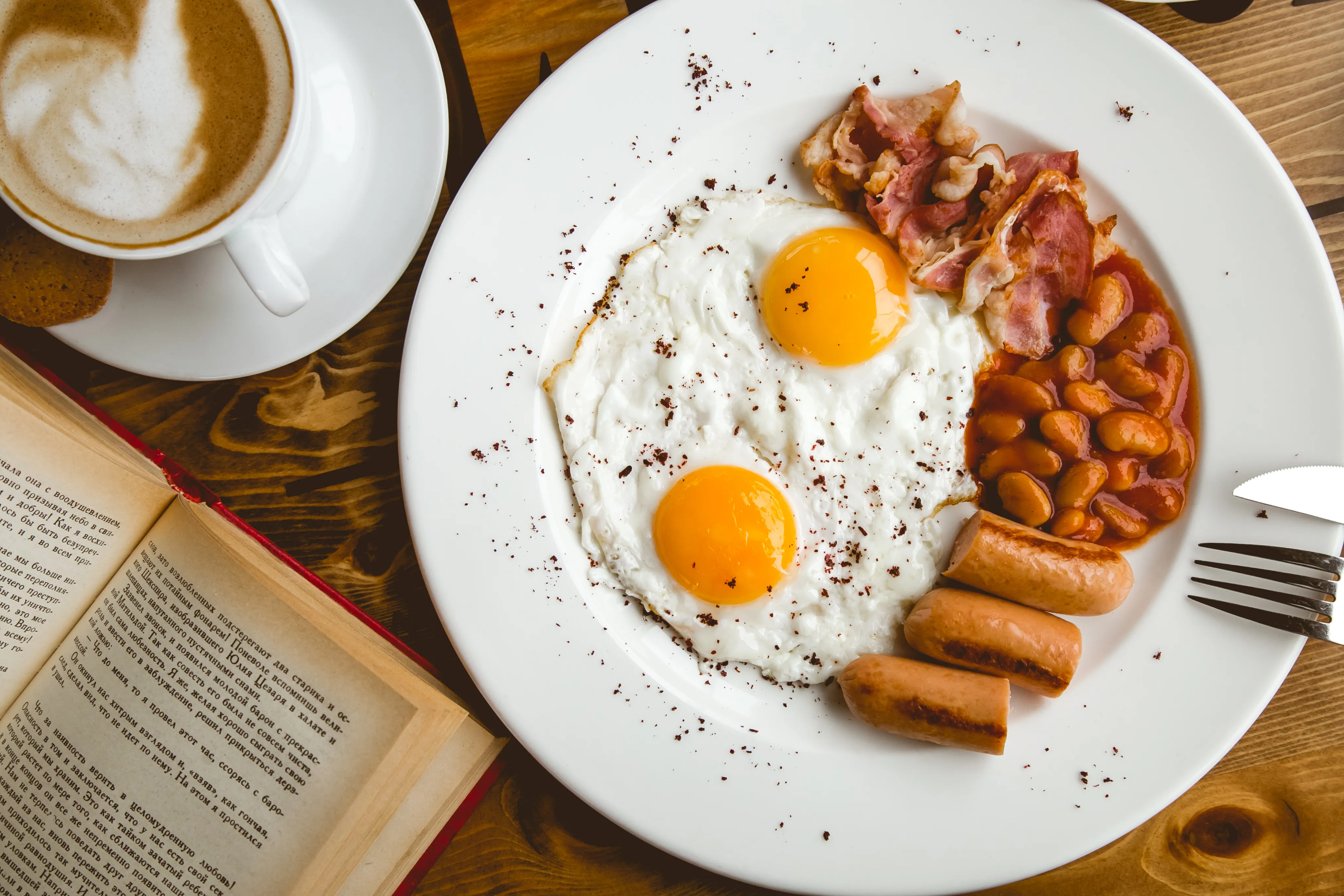 Plate with sunny side up eggs, bacon, baked beans, and sausages. A latte and open book are on a wooden table, creating a cozy breakfast scene
