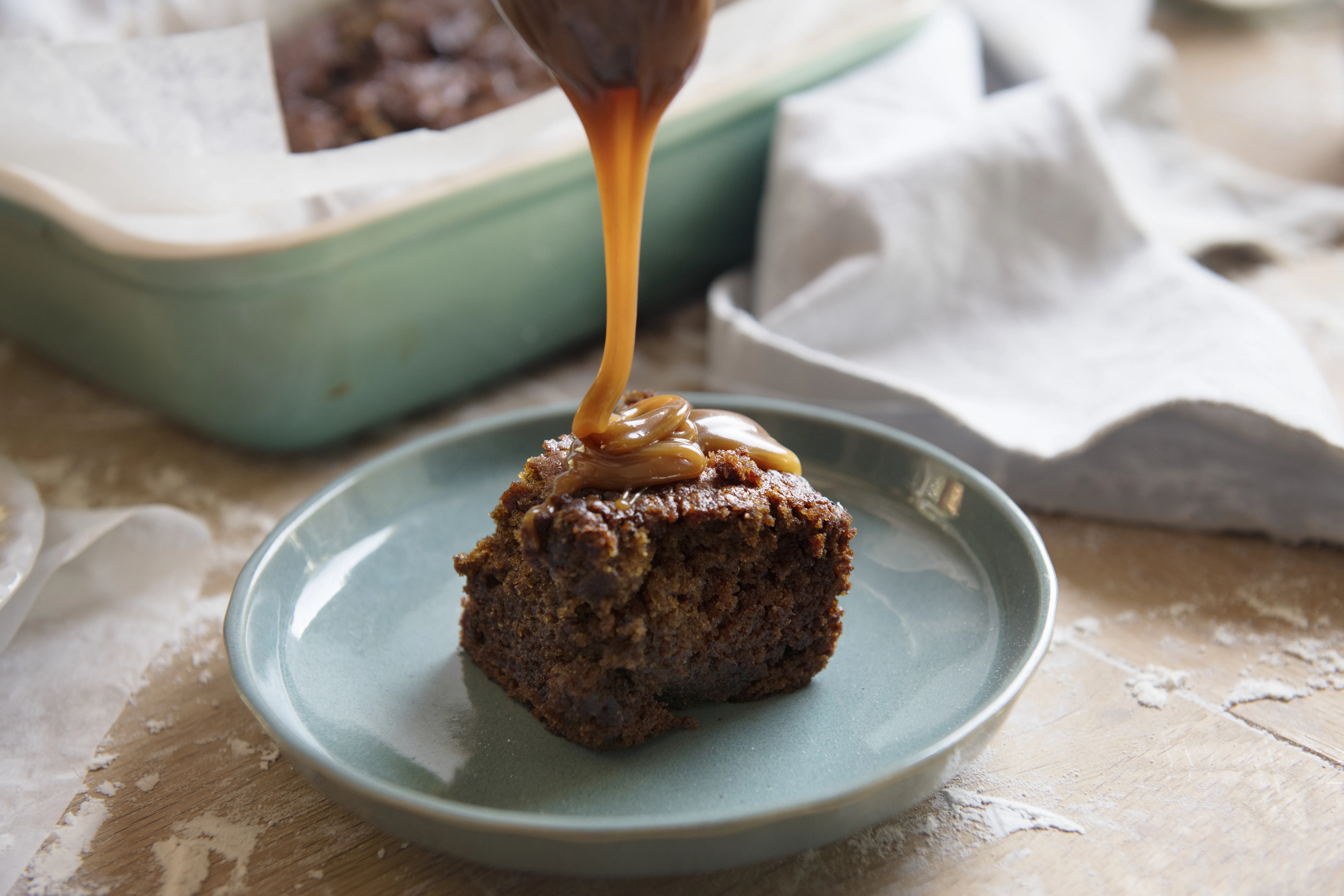 A rich, sticky toffee cake square on a teal plate is drizzled with caramel sauce, evoking warmth and indulgence. A baking dish and napkin are in the background.