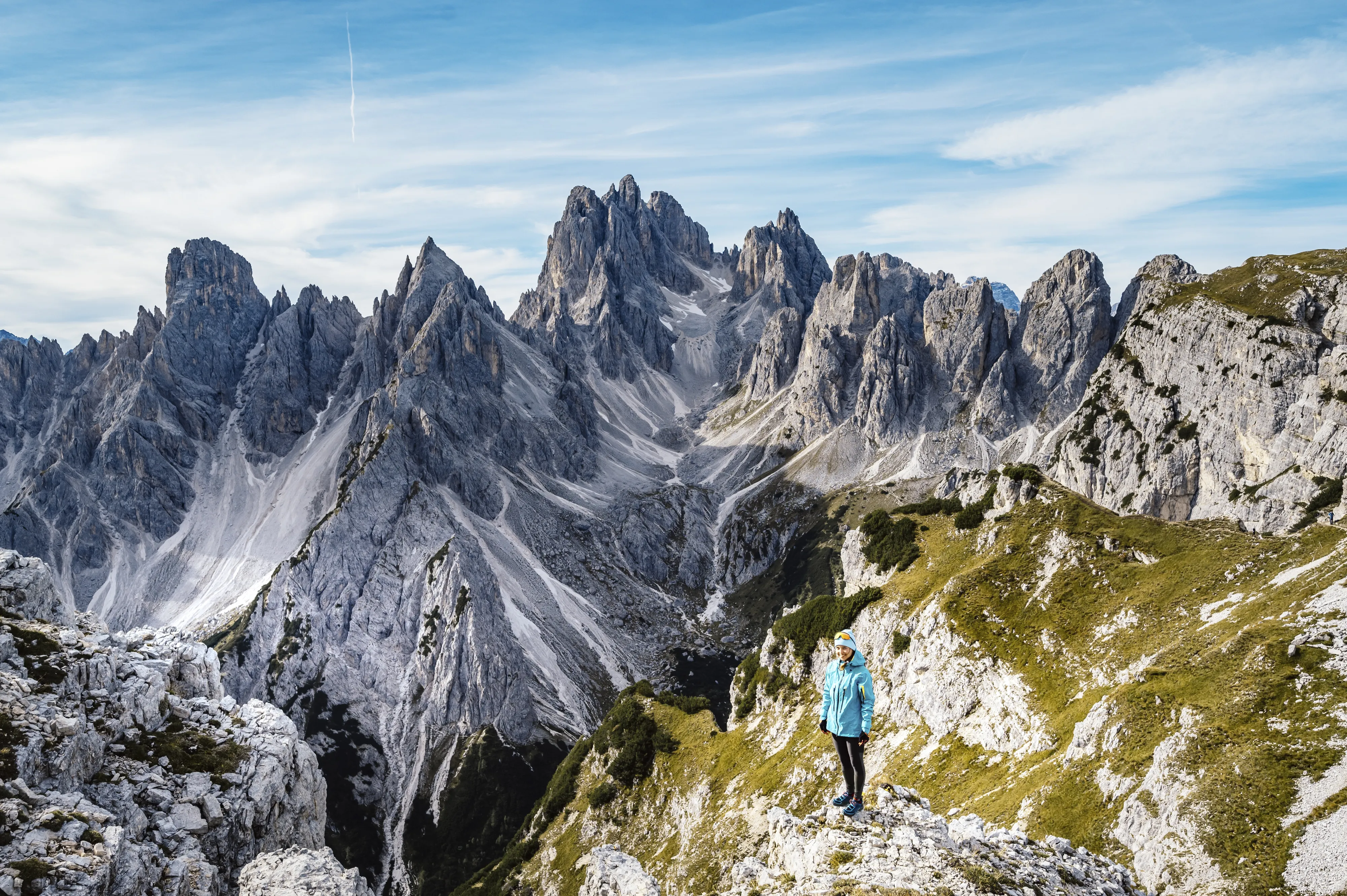 A lone hiker stands on a grassy mountain ridge overlooking the jagged Cadini di Misurina peaks.