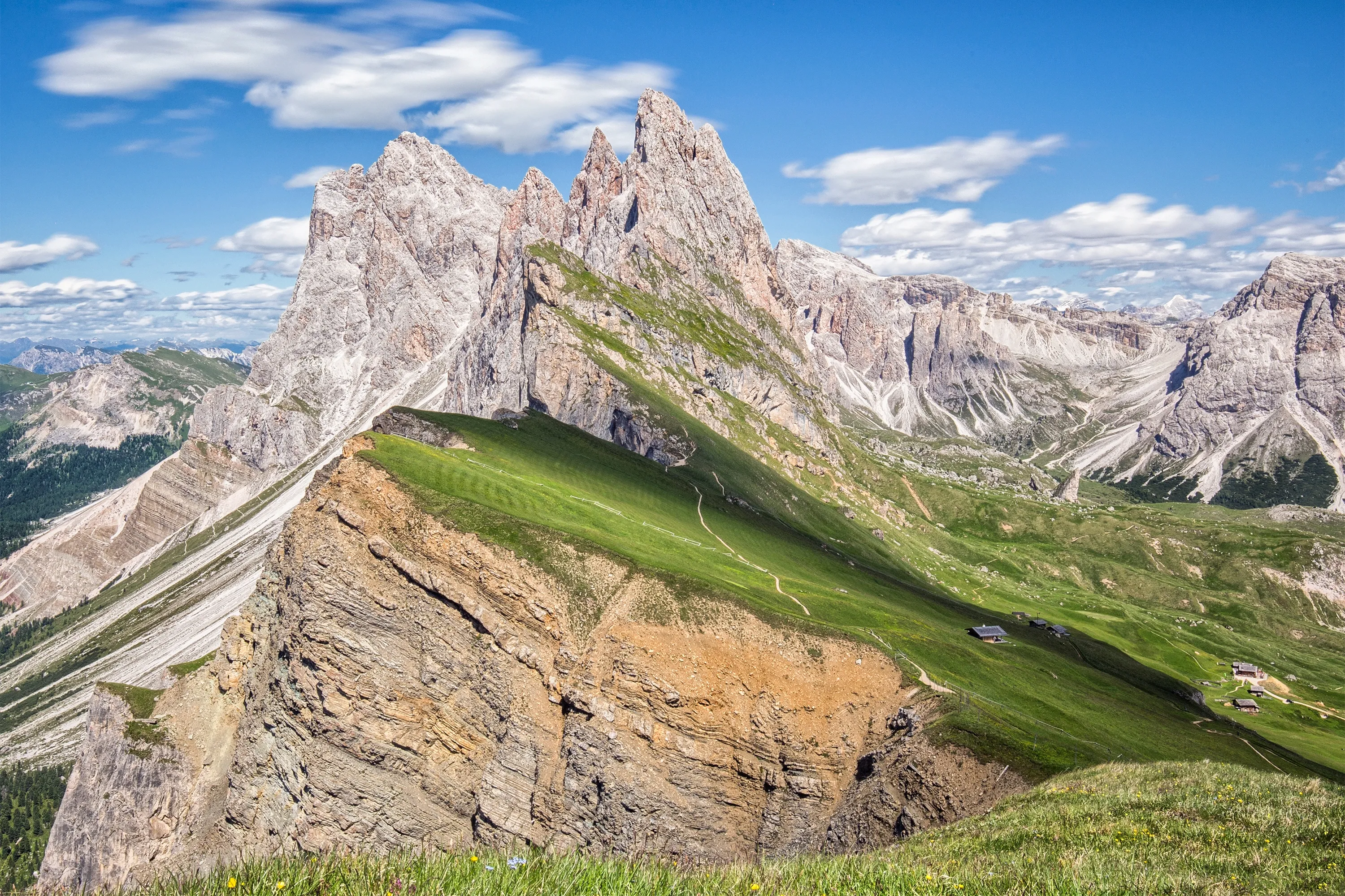 A green, sloped ridge leads up to the jagged Seceda mountain peaks under a cloudy blue sky.