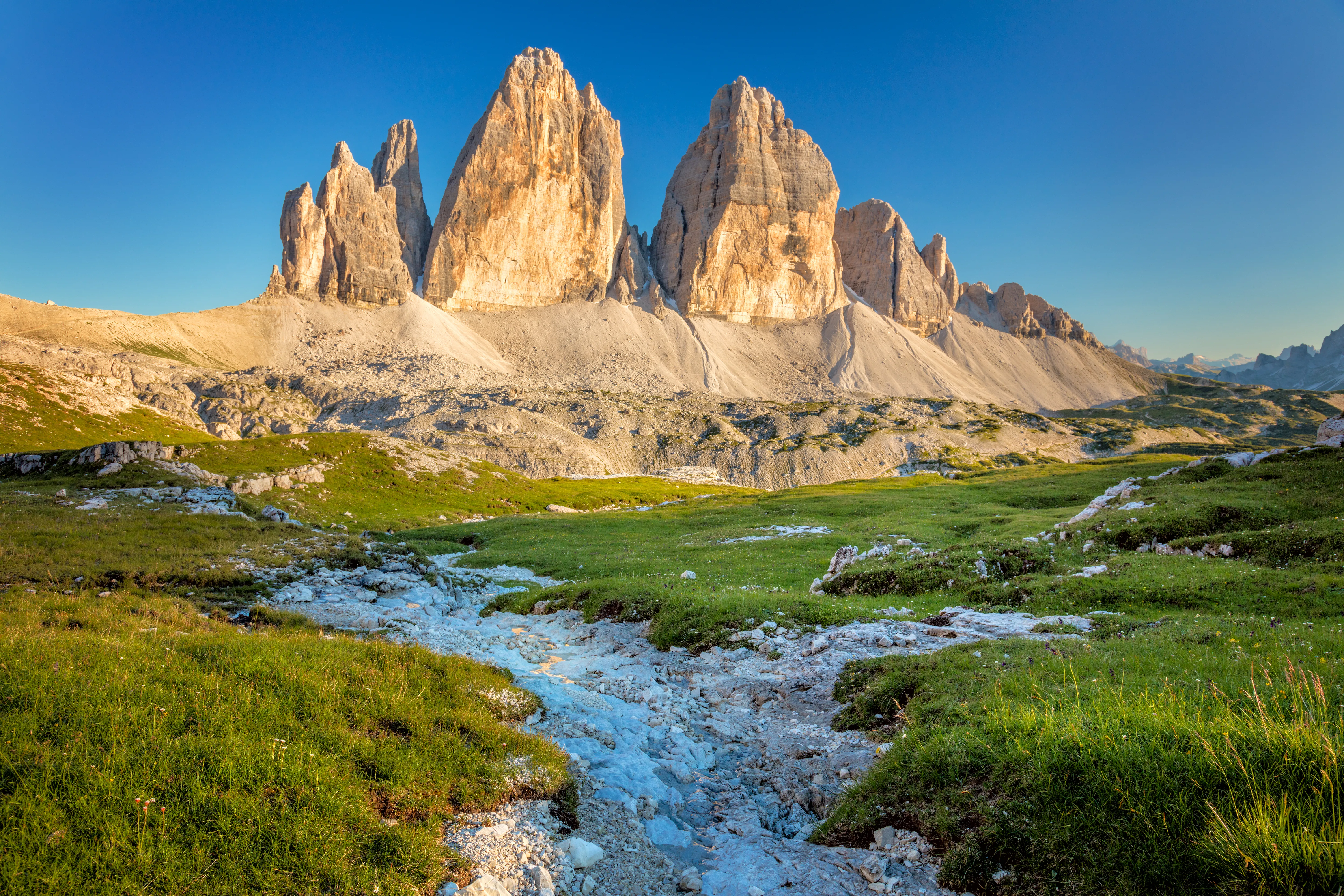 A wide-angle, eye-level shot of the iconic Tre Cime di Lavaredo (Three Peaks) in the Dolomites, Italy, illuminated by warm afternoon sunlight.