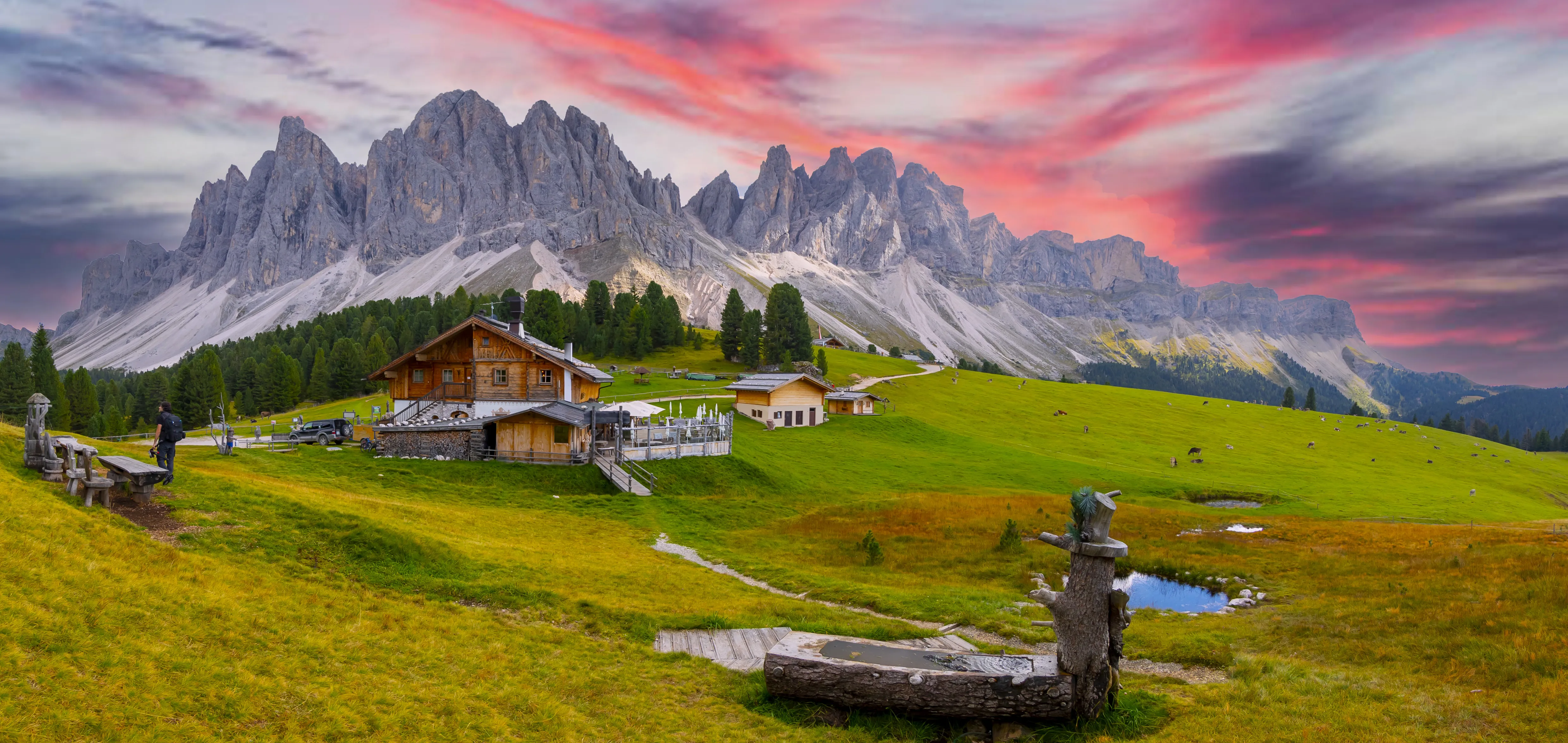 A wide-angle, eye-level panoramic shot of a rustic wooden mountain hut nestled in a rolling green alpine meadow within the Dolomites, Italy.