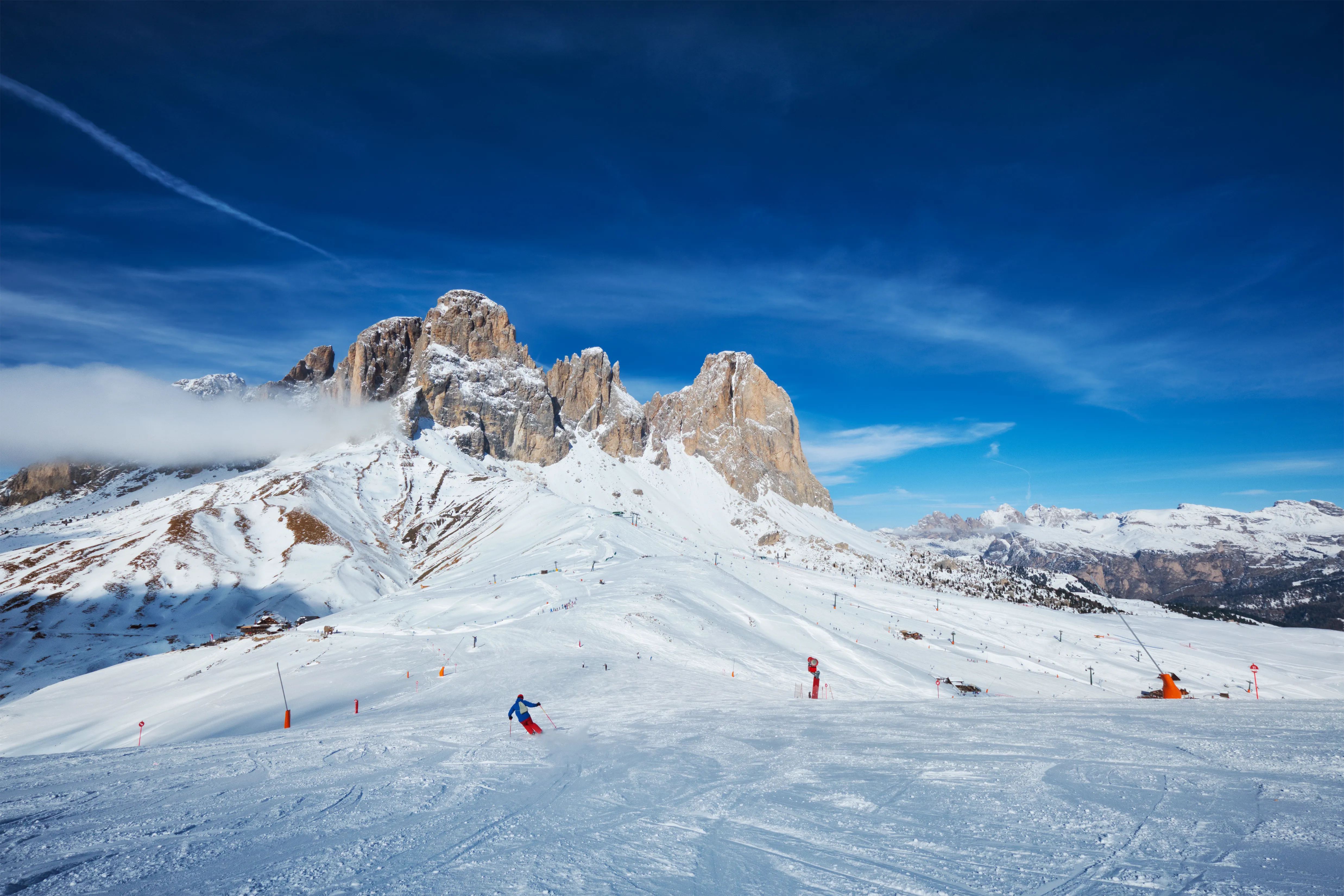 A lone skier descends a vast snowy slope in the Dolomites under a bright blue sky.